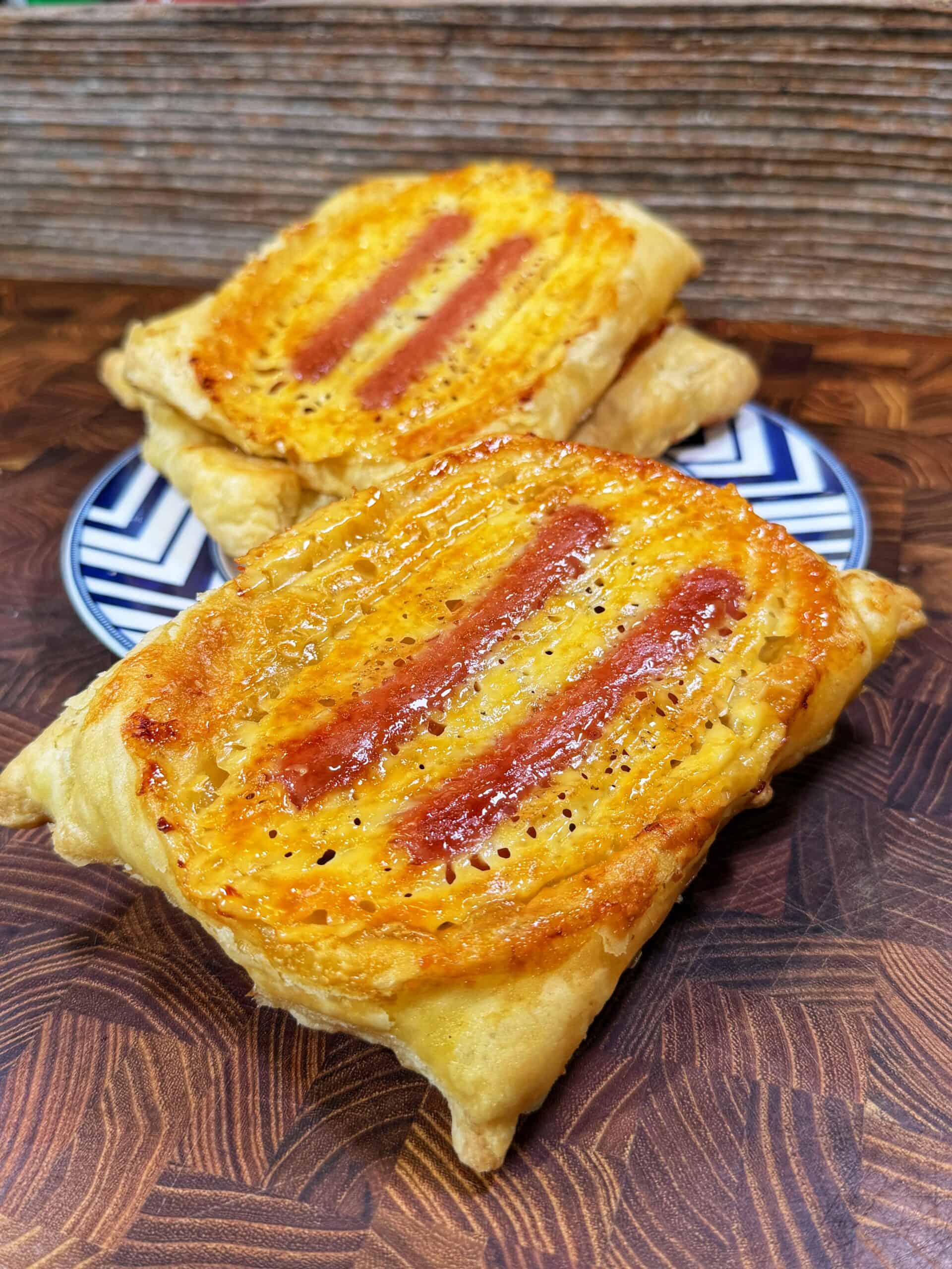 Two rectangular puff pastries with a golden, flaky crust and two reddish filling stripes on top are placed on a patterned plate and a wooden surface. A rustic wooden panel serves as the background.