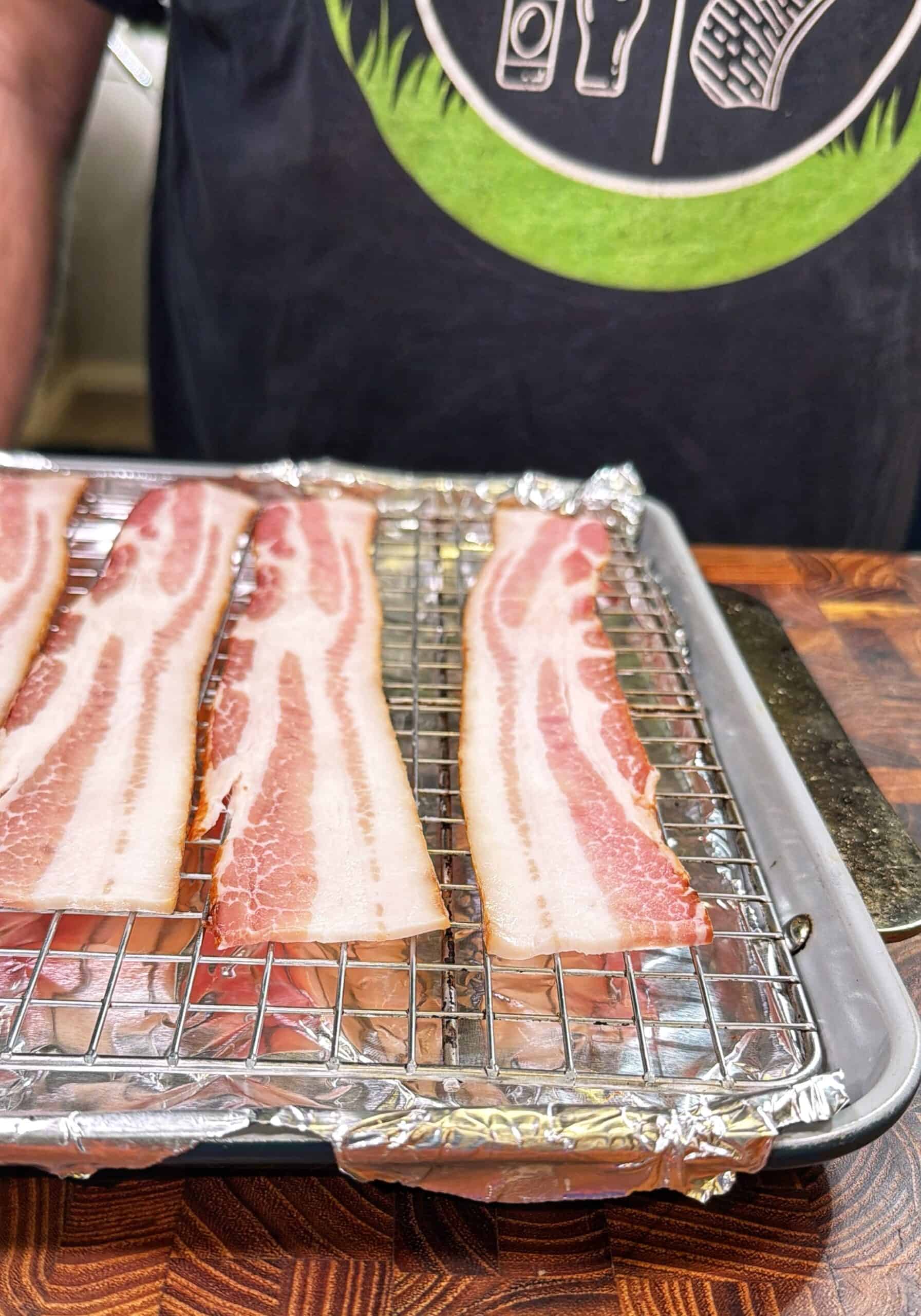 Four raw bacon strips are laid out on a wire rack over a foil-lined baking sheet. A person wearing a dark shirt with kitchen-themed graphics stands behind the tray. The scene is set on a wooden surface.