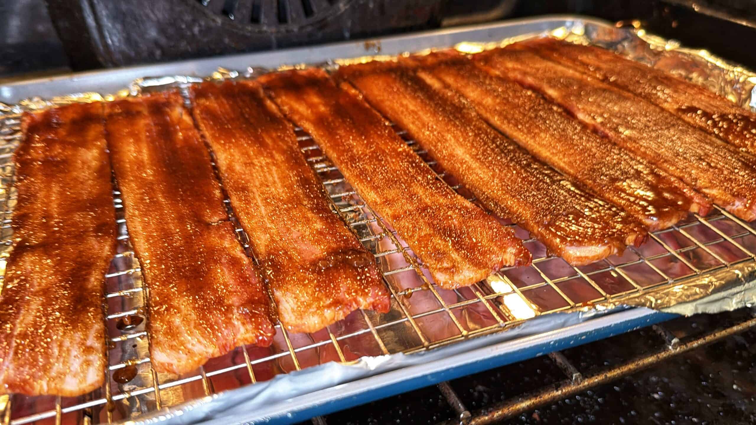 Strips of bacon coated with seasoning are arranged on a wire rack over a foil-lined baking sheet inside an oven, ready to be cooked.