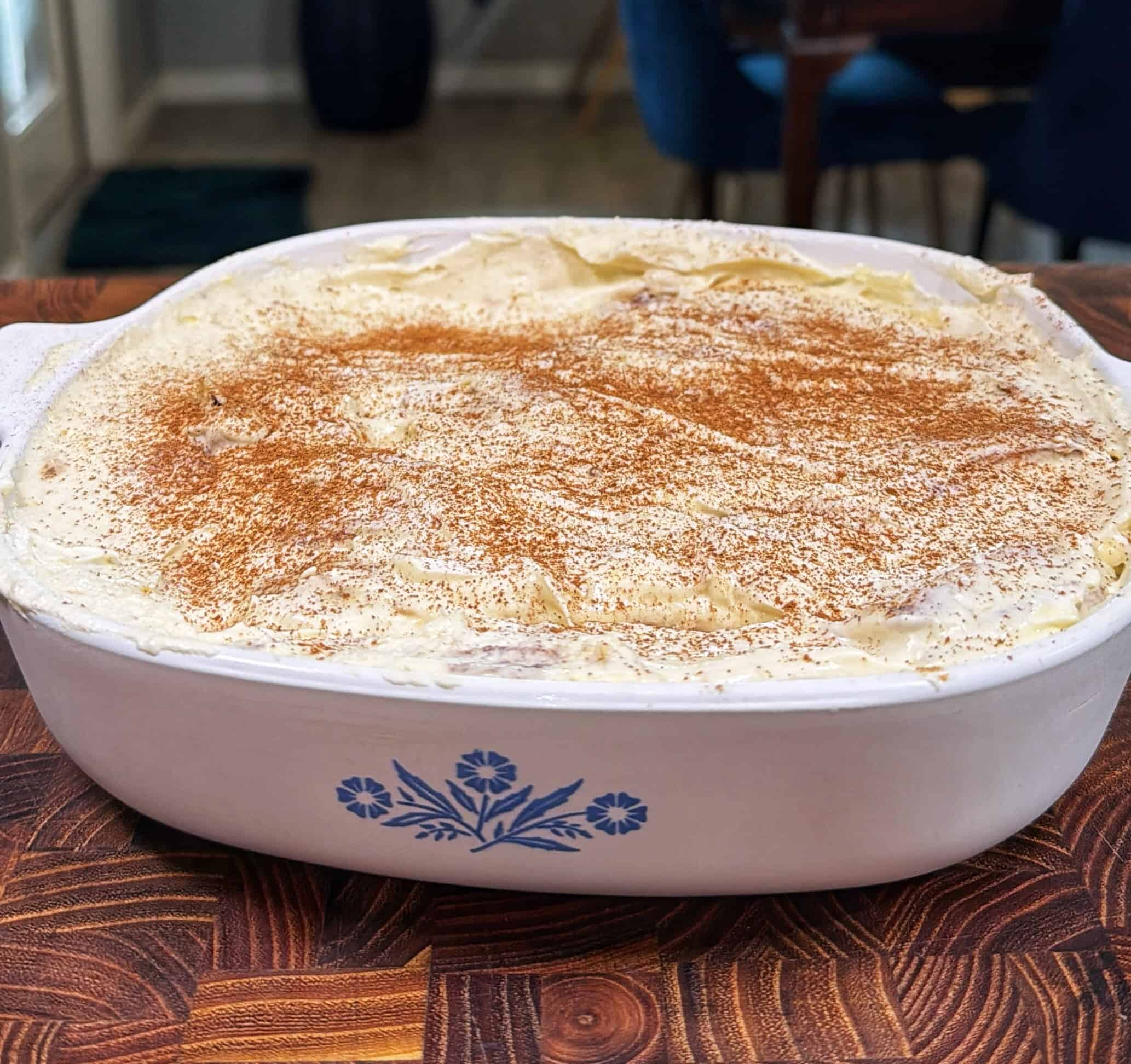 A rectangular white casserole dish with a blue floral design holds a creamy, white-topped dessert sprinkled with cinnamon, sitting on a wooden surface.