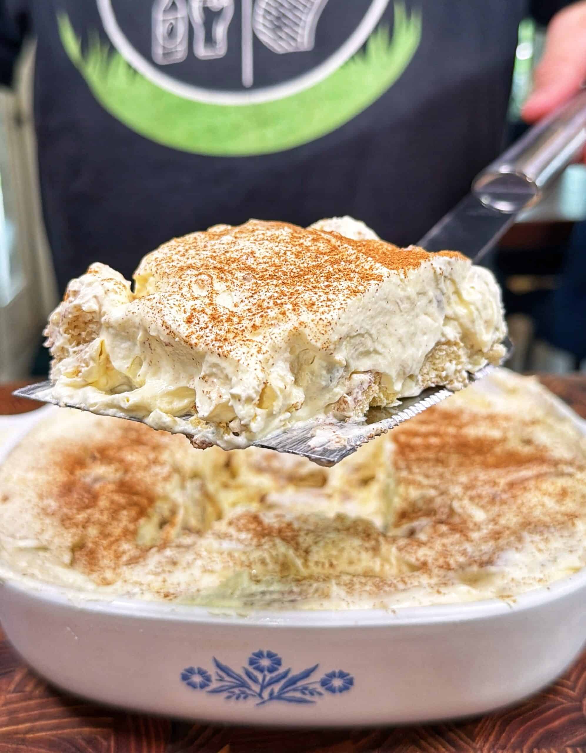 A close-up of a creamy dessert, possibly tiramisu or banana pudding, being served from a white dish with a blue floral design. The dessert is topped with powdered cinnamon and has a rich, fluffy texture.