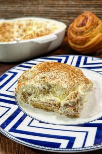 A slice of creamy dessert topped with cinnamon sits on a blue and white patterned plate; behind it are a baking dish with more dessert and a round, swirled roll on a wooden surface.