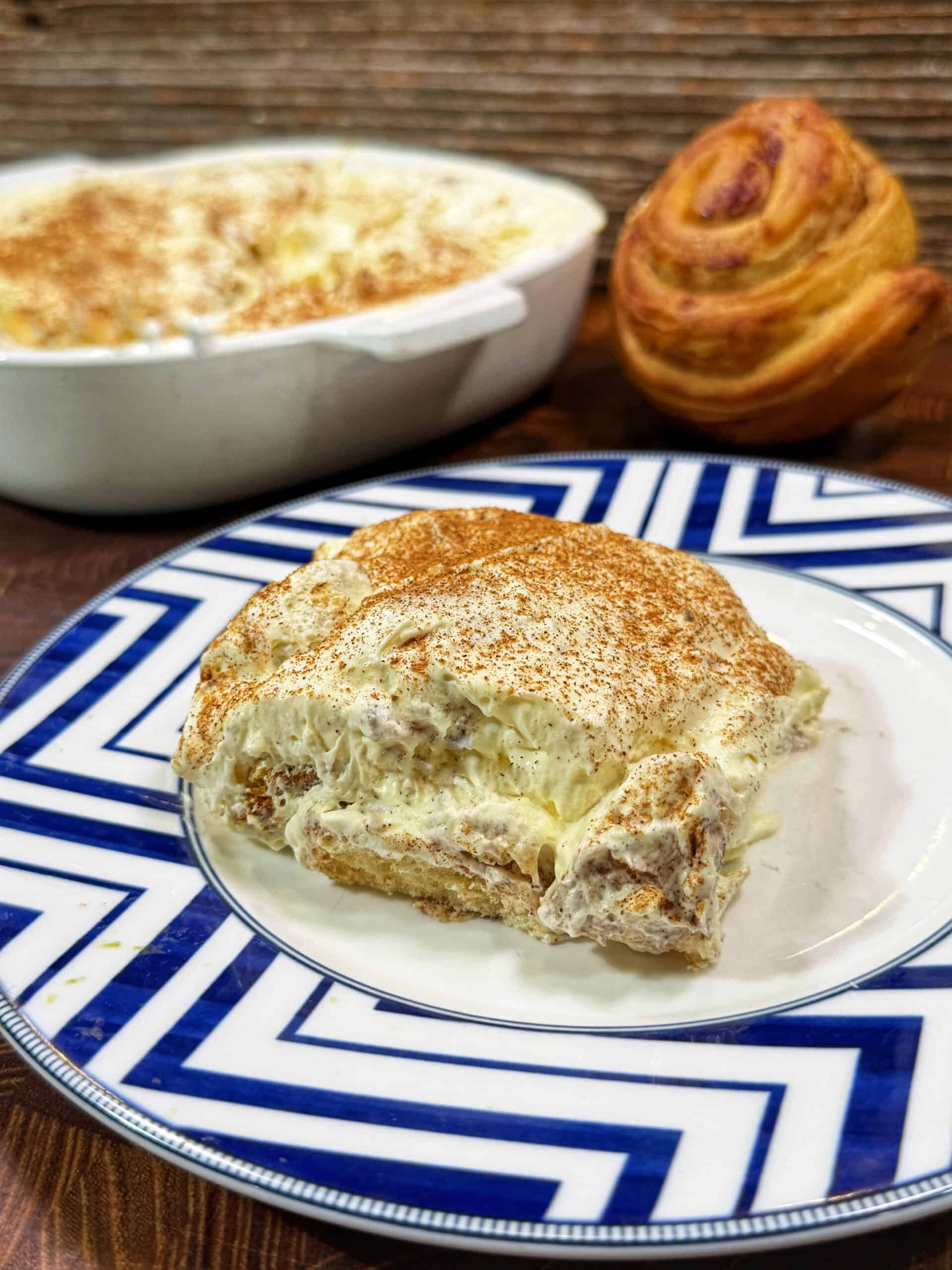 A slice of creamy dessert topped with cinnamon sits on a blue and white patterned plate; behind it are a baking dish with more dessert and a round, swirled roll on a wooden surface.