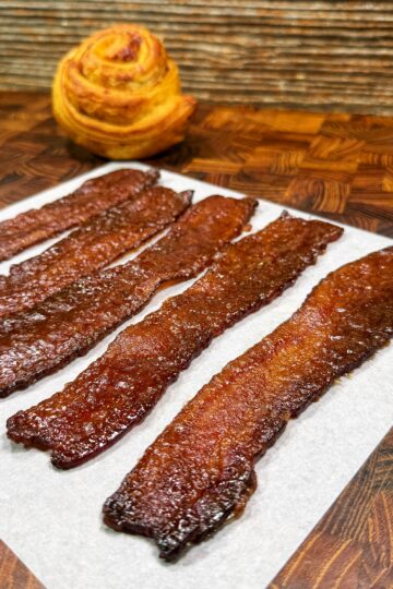 Four strips of crispy candied bacon are laid out on a piece of parchment paper atop a wooden surface, with a round, spiral pastry in the background.