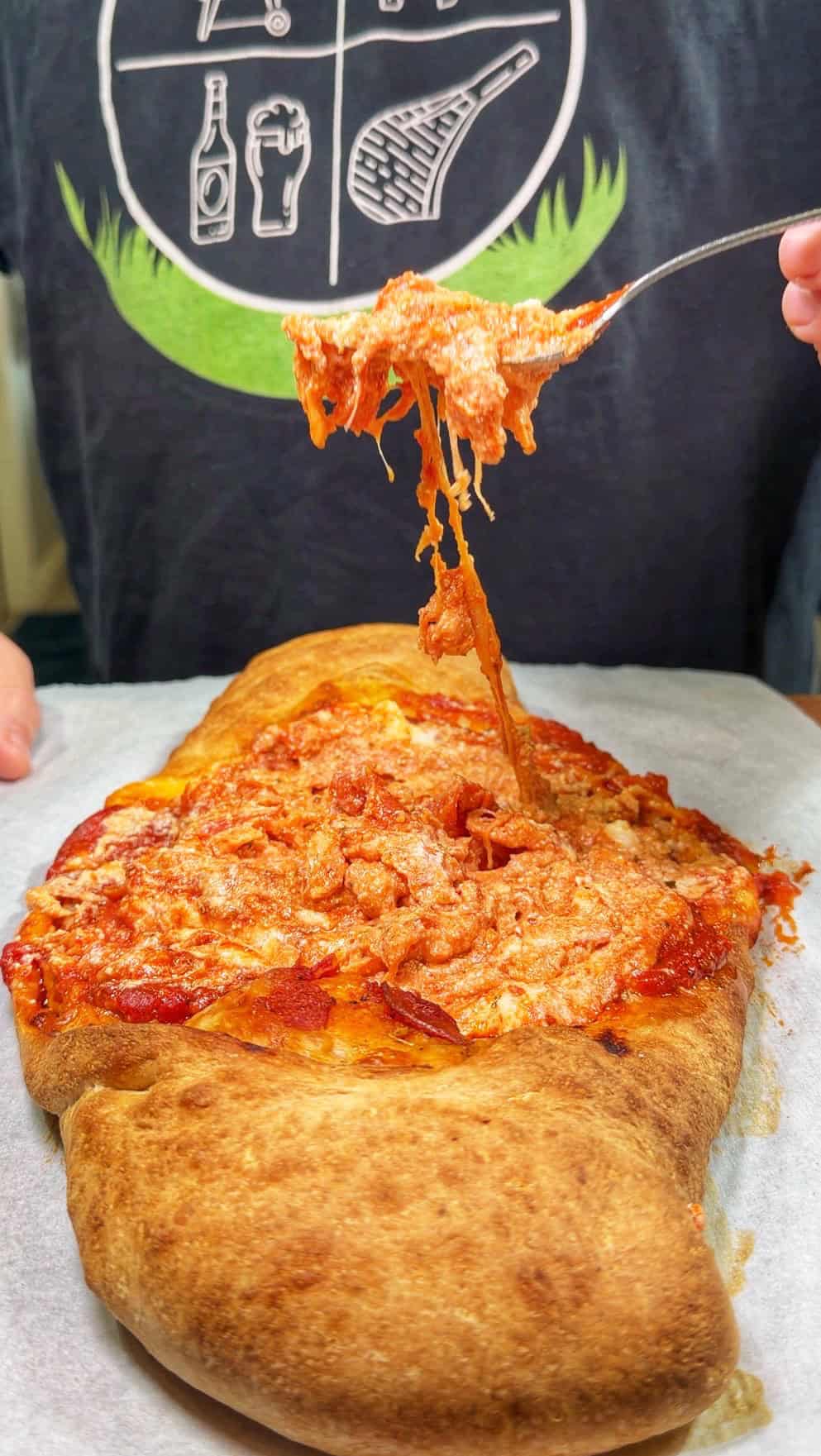 A person lifts a forkful of cheesy, saucy filling from an oval-shaped calzone on parchment paper, with melted cheese stretching upward. The person’s T-shirt features food and drink icons.