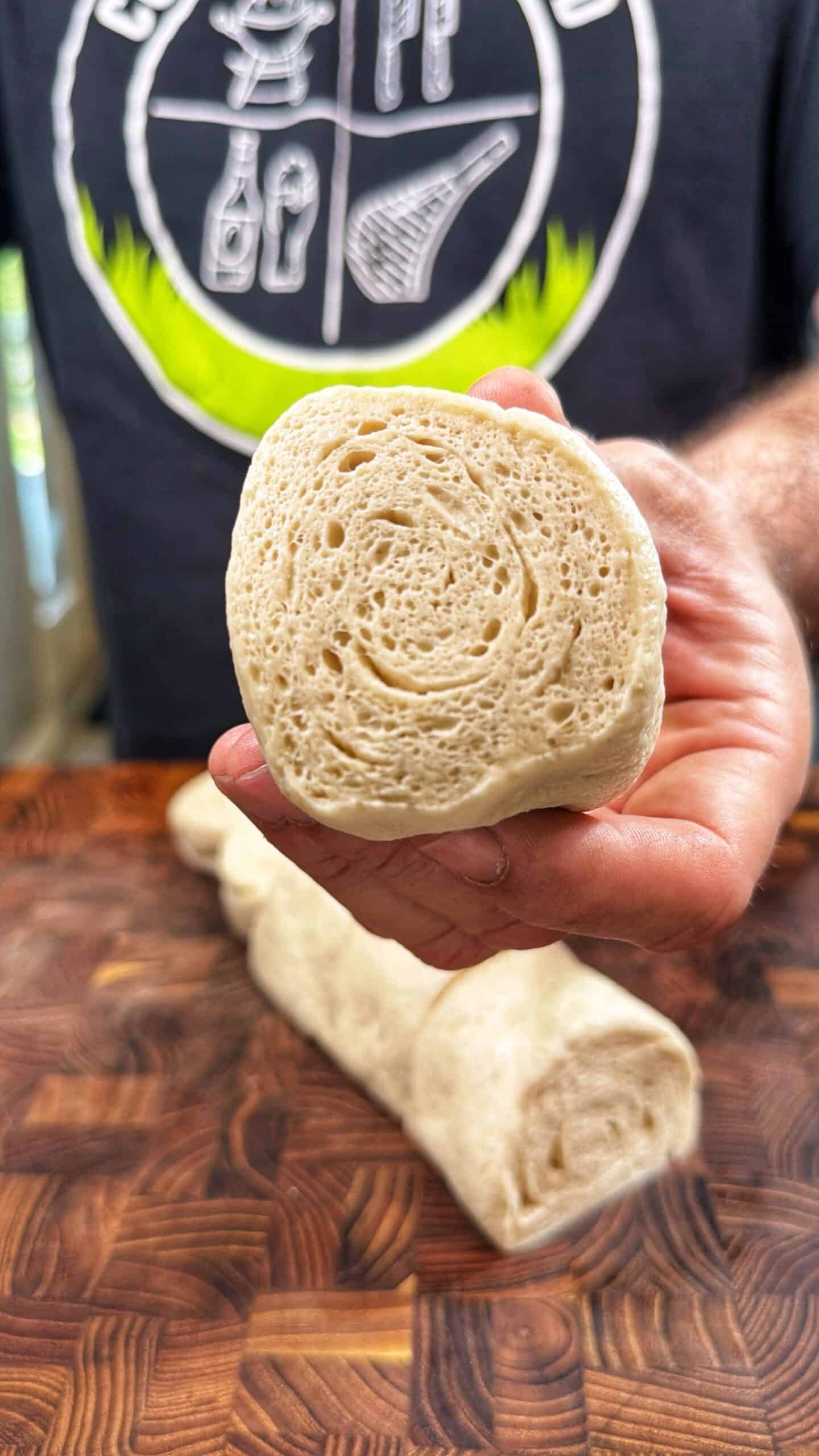 A person holds a halved piece of rolled dough, showing its airy, bubbly interior. Another piece of dough rests on a wooden cutting board. The person wears a dark shirt with a white illustrated design.