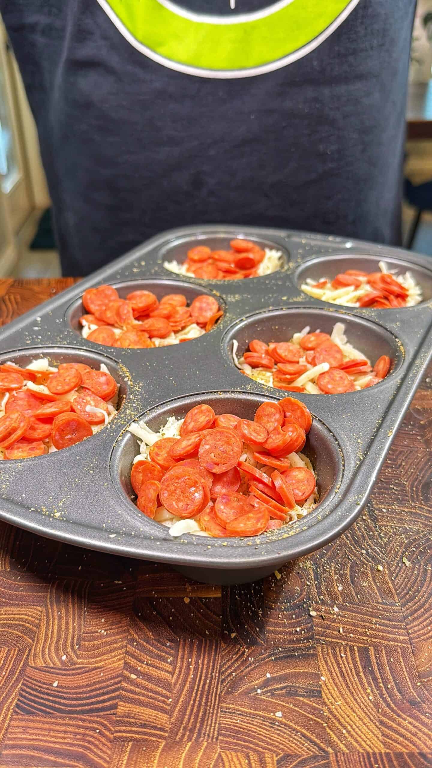 A muffin tin filled with shredded cheese and small pepperoni slices, sitting on a wooden countertop. Someone wearing a dark shirt with a green circular logo holds the tin.