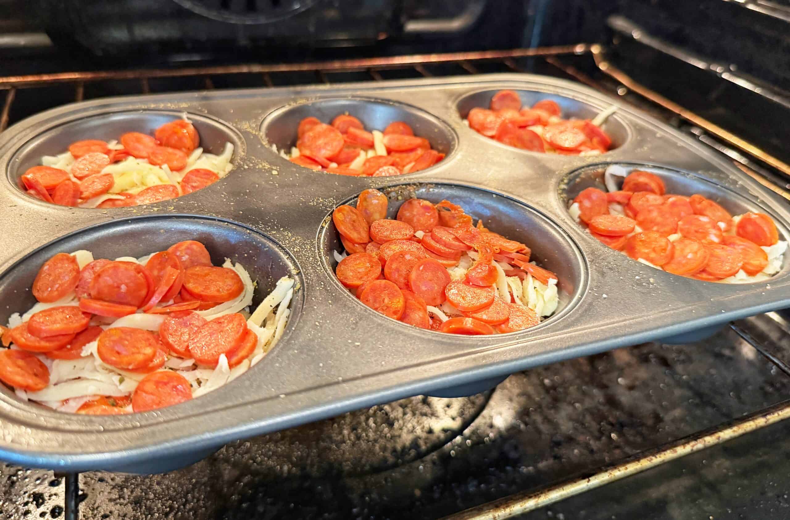 A muffin tin filled with mini pizza ingredients, including shredded cheese and sliced pepperoni, is baking in an oven.