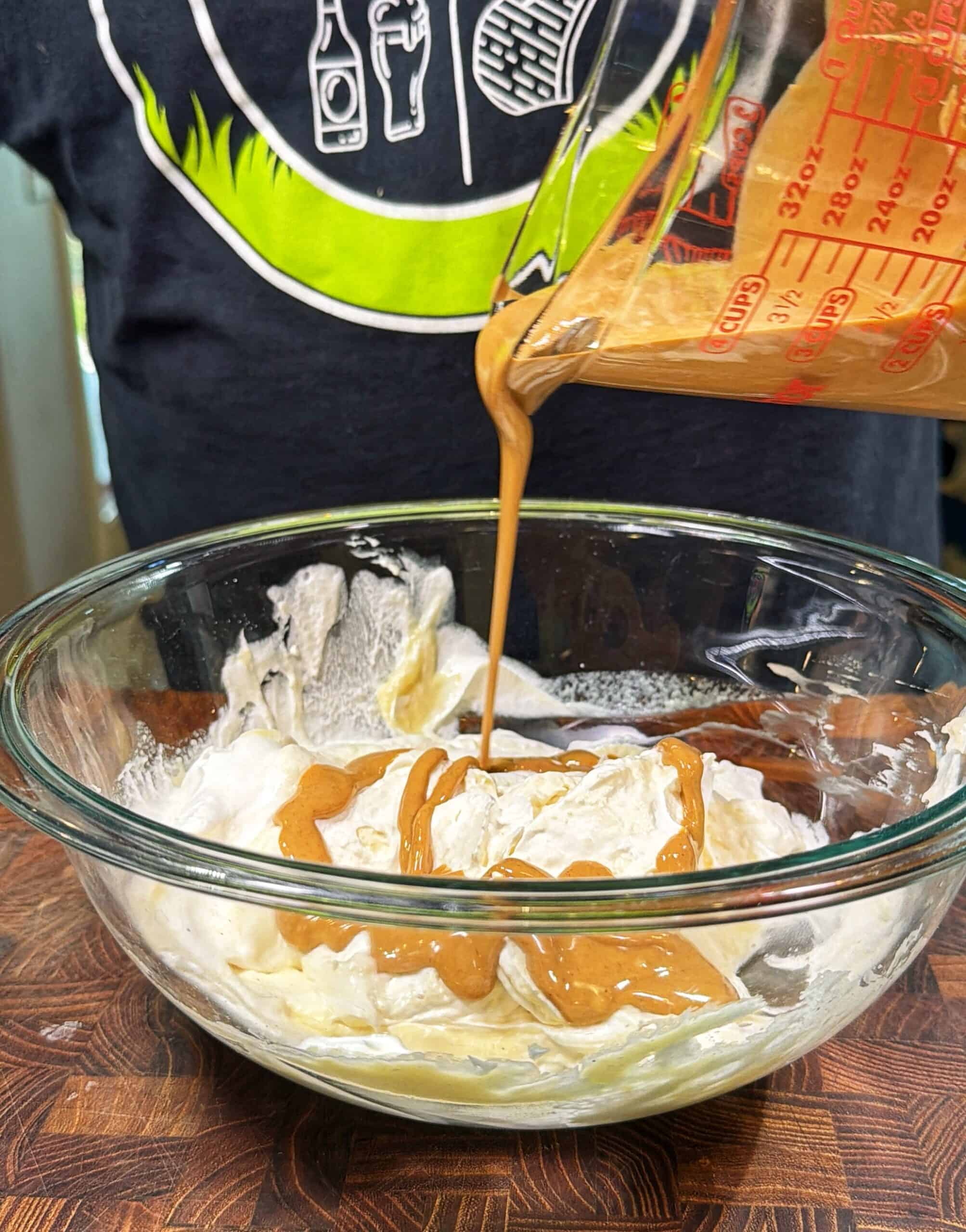 A person pours a light brown liquid from a measuring cup into a glass bowl containing a white creamy mixture, all set on a wooden surface.