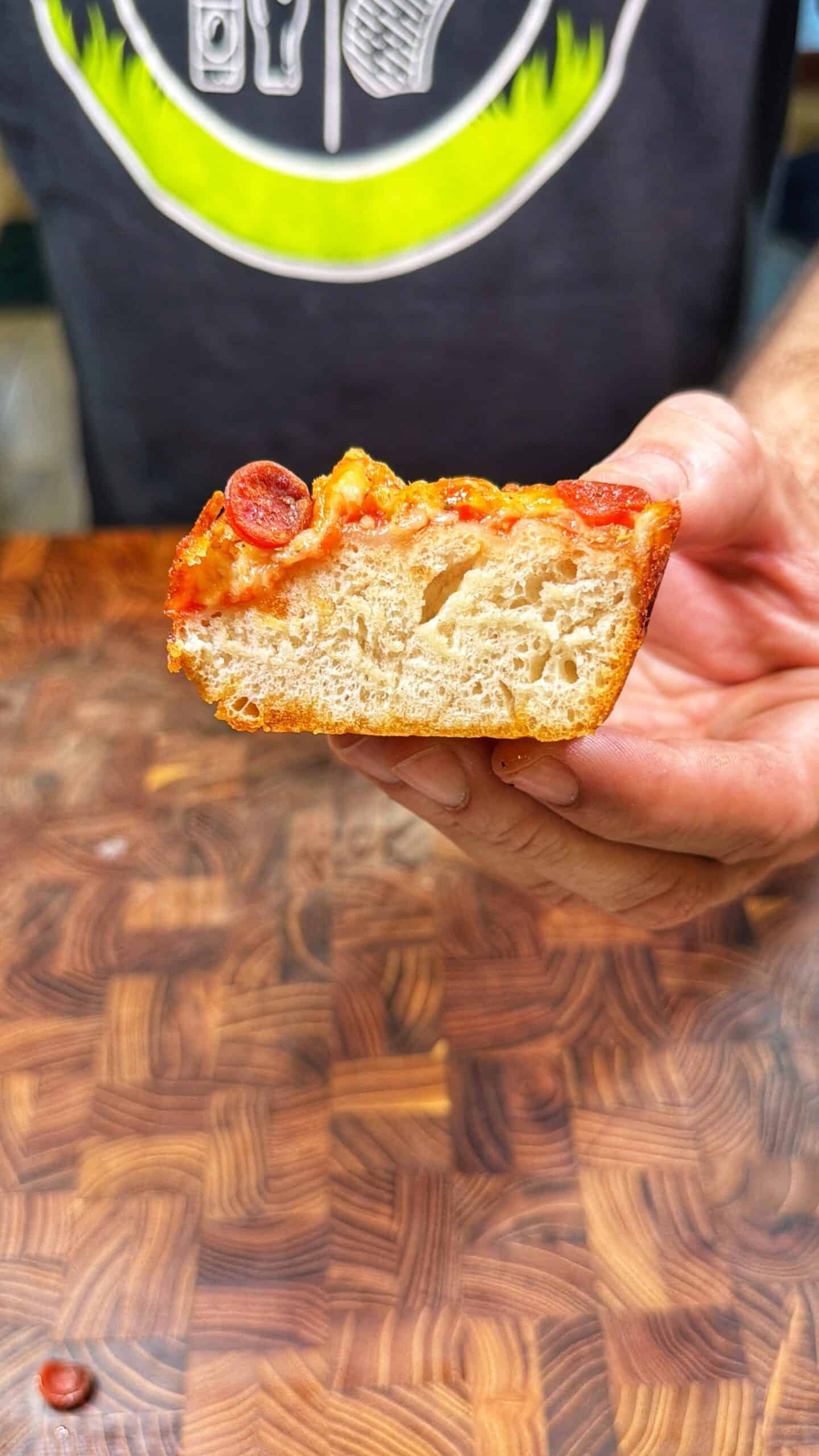 A hand holds up a thick slice of pizza showing its airy, soft crust, melted cheese, tomato sauce, and a small slice of pepperoni on top, with a wooden surface and a person wearing a dark shirt in the background.
