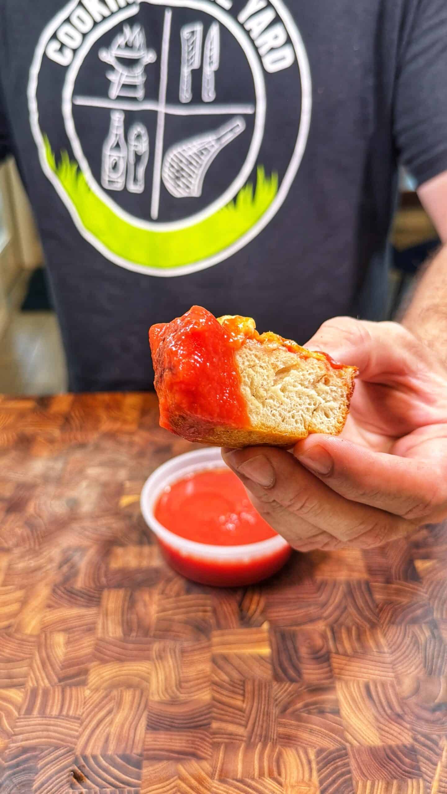 A person holds a piece of bread with tomato sauce in front of them, with a small bowl of tomato sauce on a wooden surface below. The person wears a dark shirt with a cooking-themed logo.