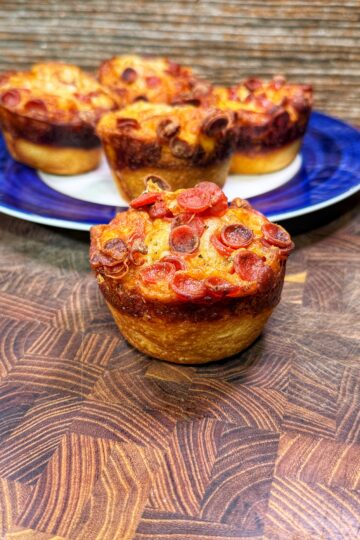 A close-up of a pepperoni pizza muffin on a wooden surface, with more pizza muffins arranged on a blue and white plate in the background.