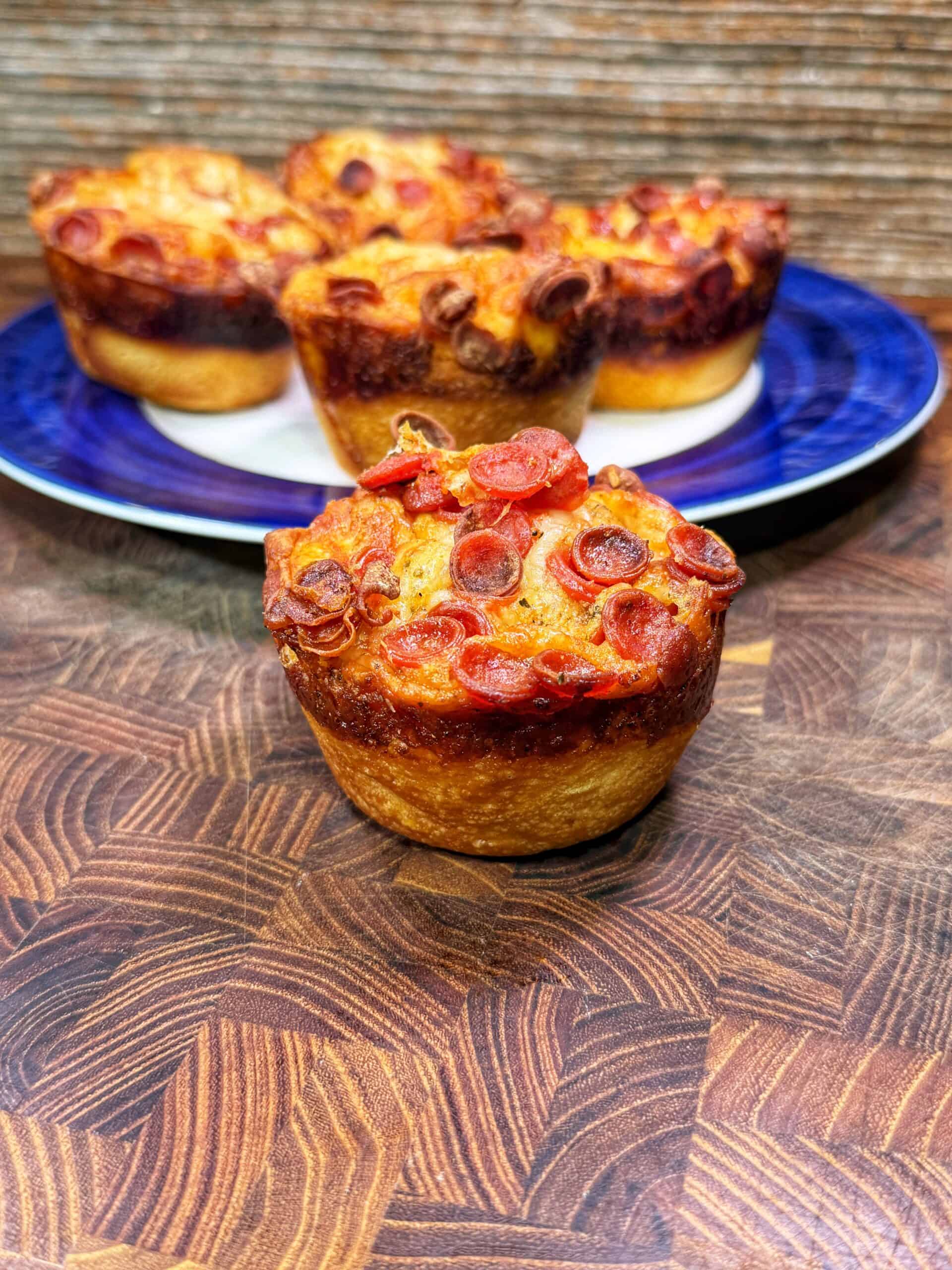 A close-up of a pepperoni pizza muffin on a wooden surface, with more pizza muffins arranged on a blue and white plate in the background.
