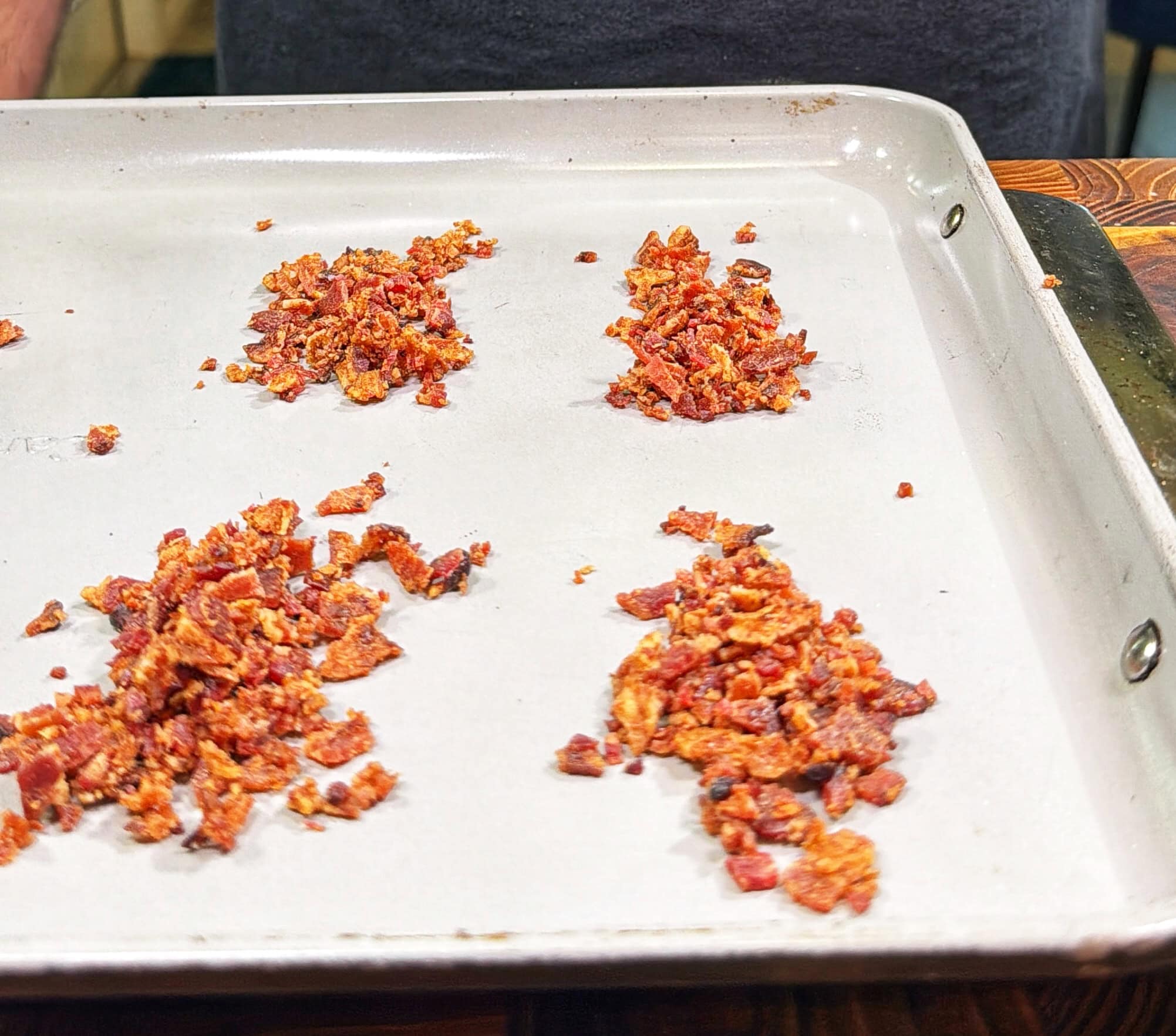 Four small piles of finely chopped sun-dried tomatoes are arranged on a large, light-colored baking sheet, with a person standing in the background.