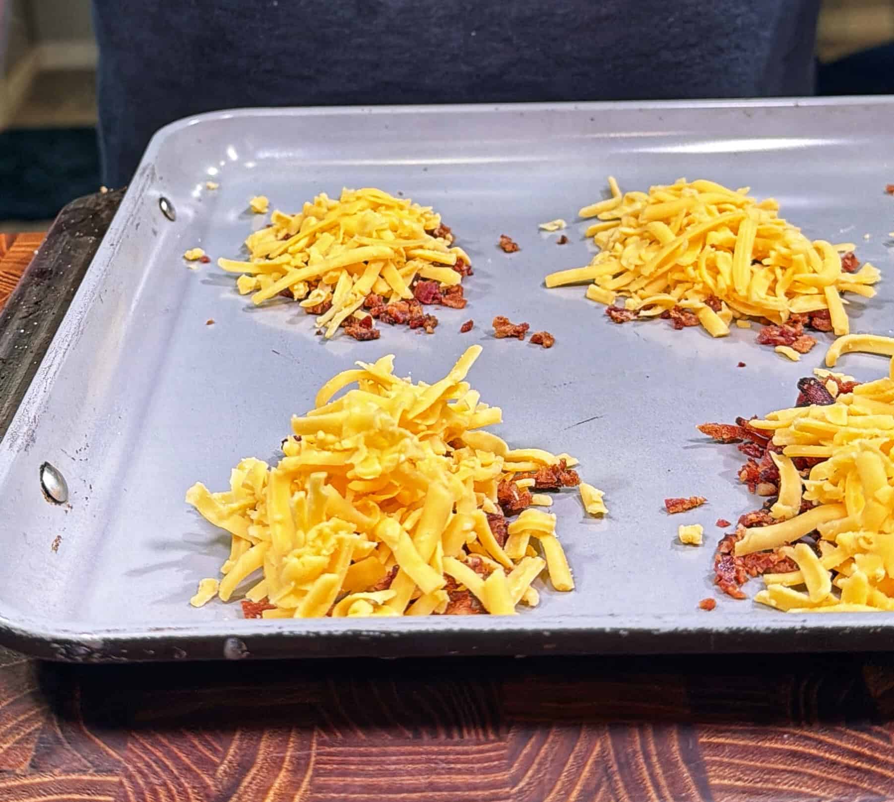 Six small piles of shredded cheddar cheese and bits of bacon are arranged on a metal baking sheet, ready to be baked. The baking sheet sits on a wooden surface, with a person partially visible in the background.