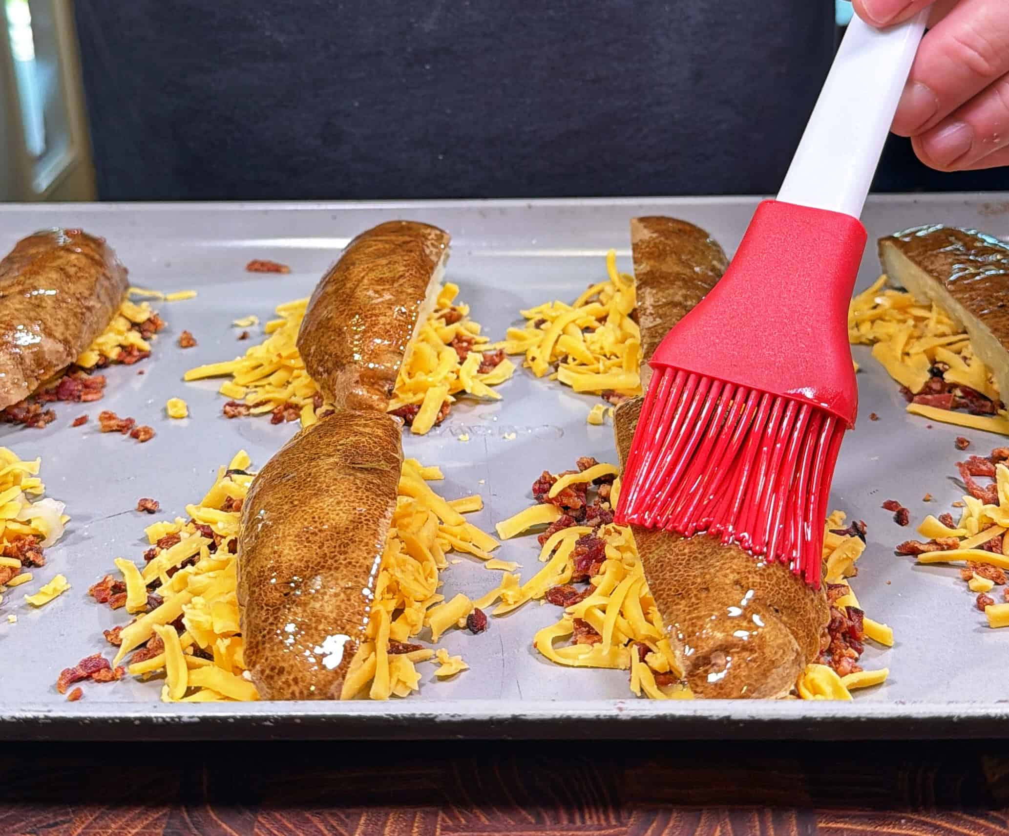 A hand uses a red pastry brush to apply butter to split baked potatoes filled with shredded cheddar cheese and bacon bits on a baking sheet.