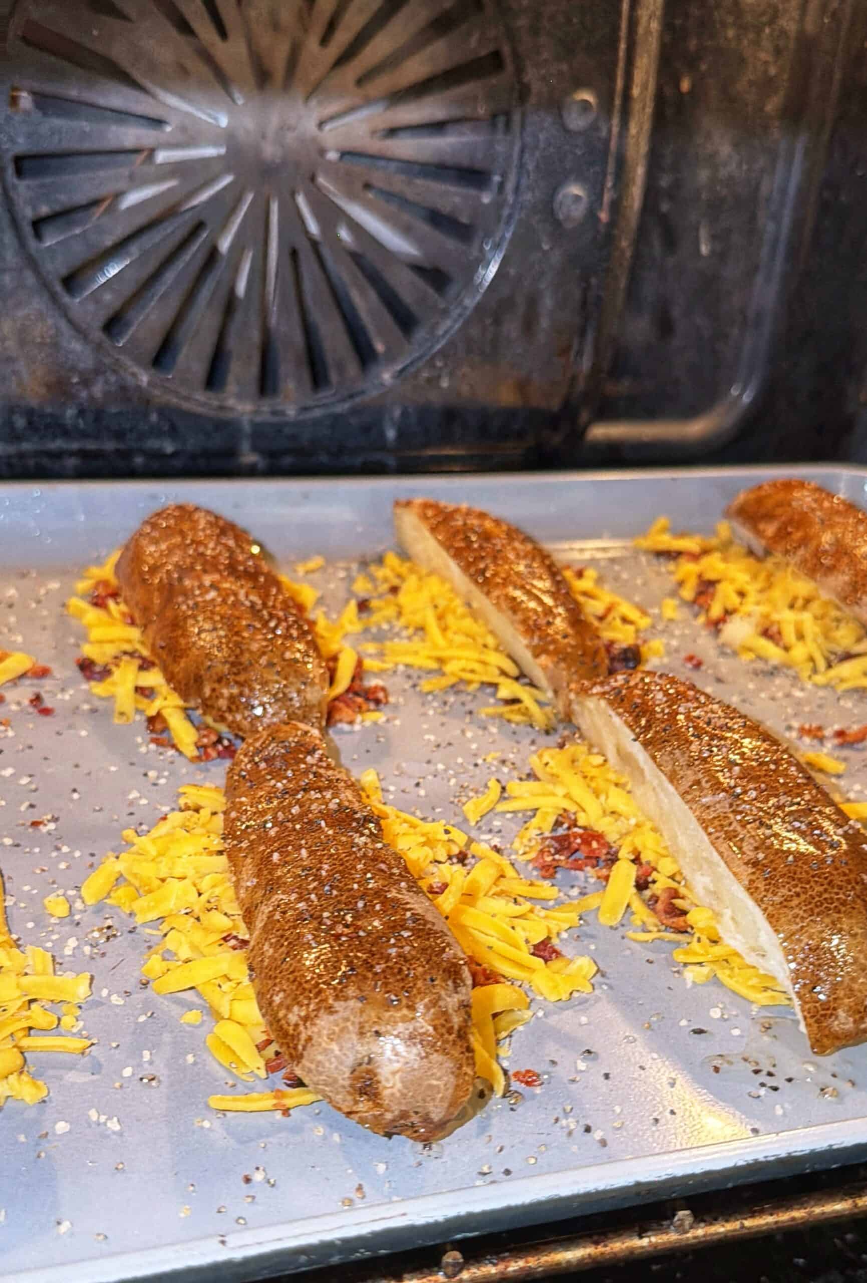 Potatoes sliced open and topped with shredded cheddar cheese and seasonings are baking on a tray inside an oven. Some cheese is melted on the tray. The oven’s interior is visible in the background.