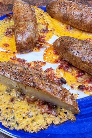 Four baked potatoes and one baked potato half are on a blue plate, surrounded by crispy melted cheese and bacon that has spread out onto the plate and hardened into a lacy, golden layer.