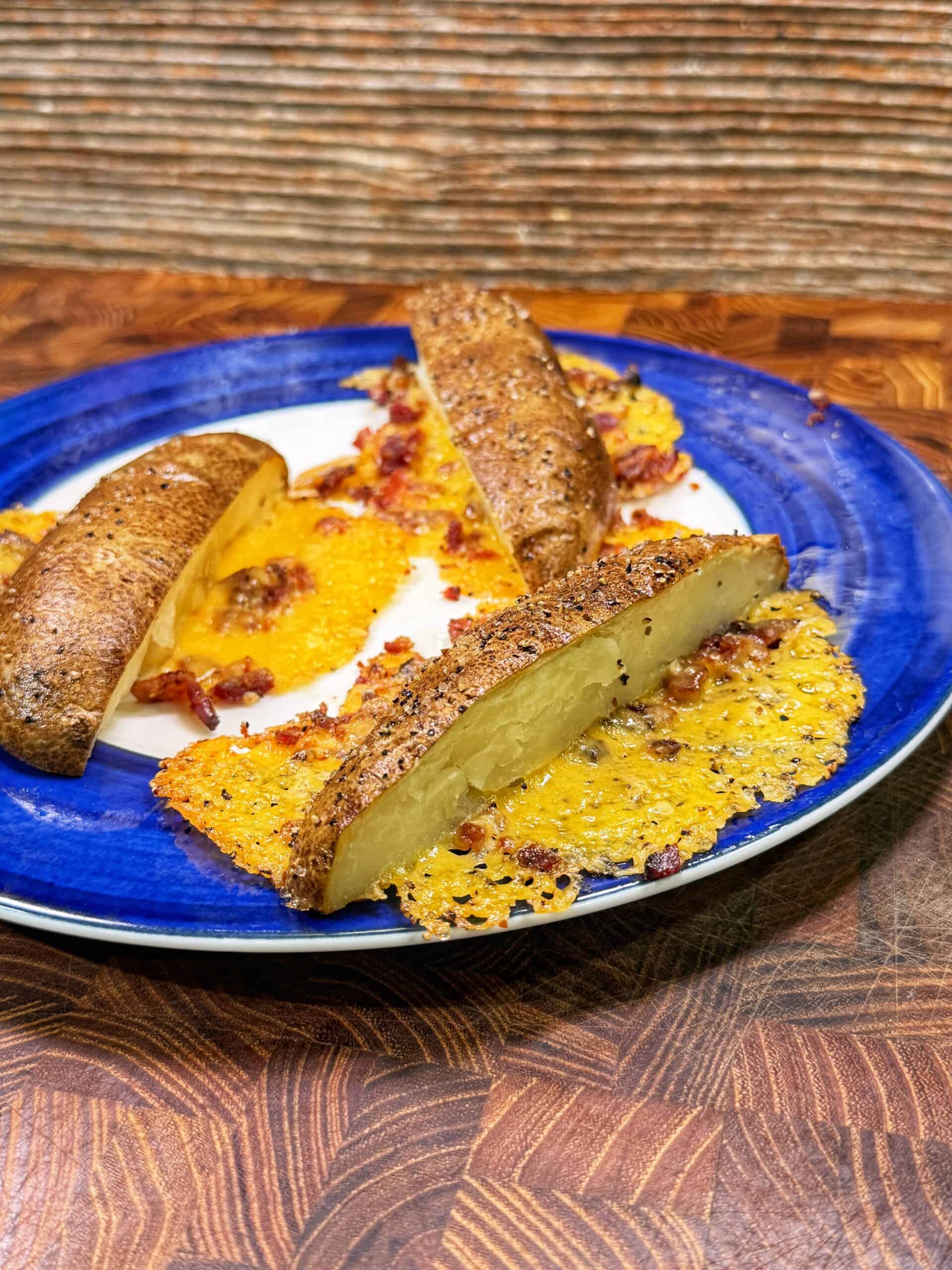 Three potato wedges on a blue and white plate, topped with crispy melted cheese and bacon bits. The plate sits on a wooden surface with a textured background.