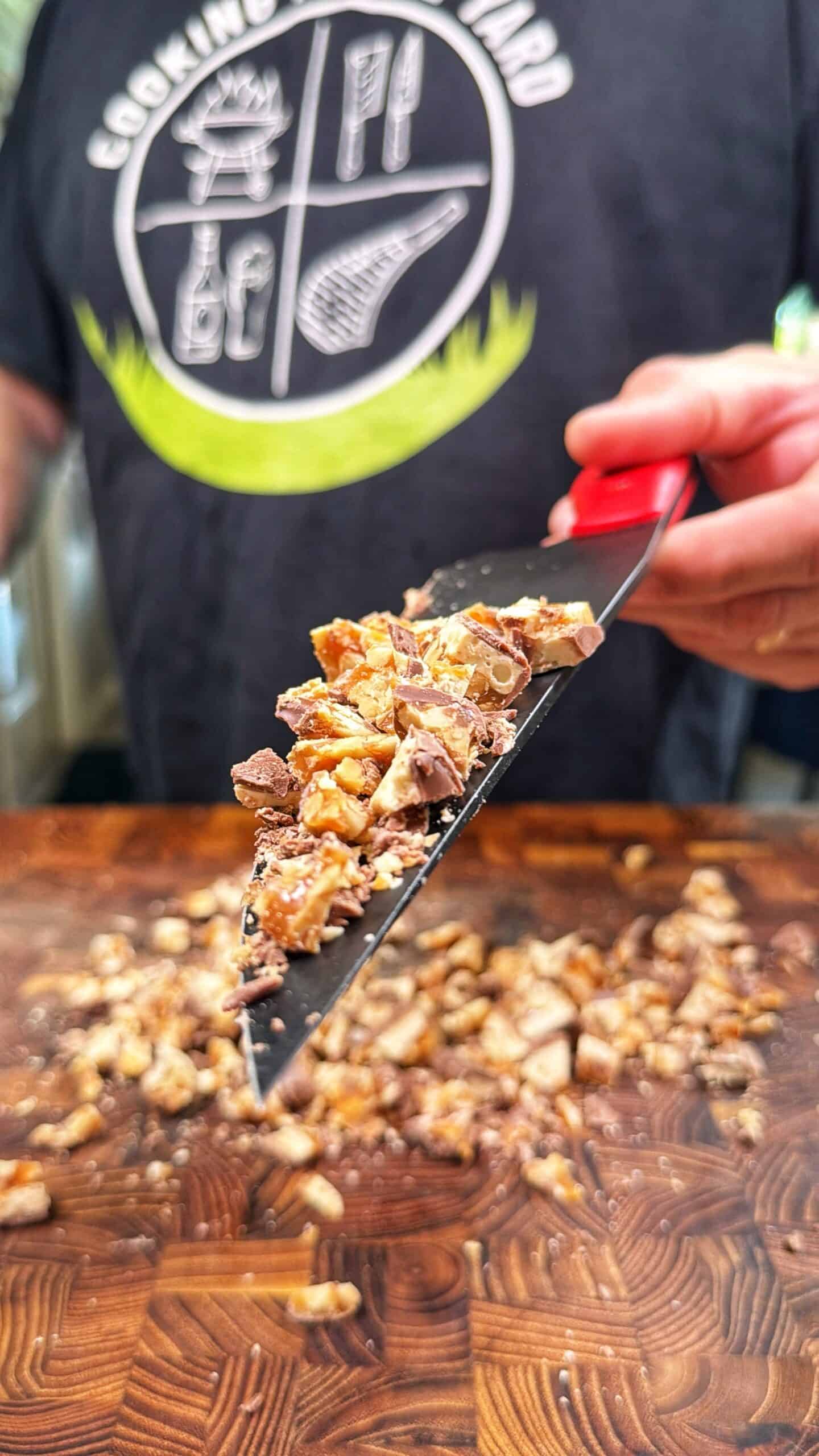 A person holds a red-handled spatula with chopped snickers bar over a wooden cutting board, while more candy is scattered on the board below. The person’s shirt has a cooking-themed graphic.
