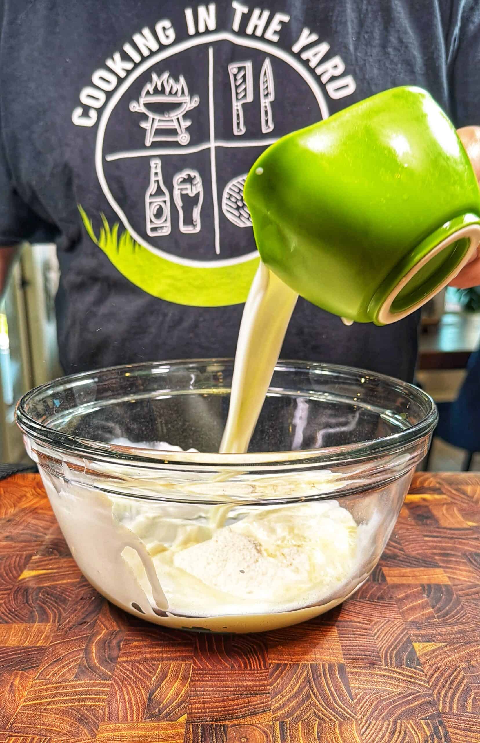 A person pours a thick, creamy liquid from a green cup into a glass bowl on a wooden surface. The person wears a shirt with Cooking in the Yard and food-related graphics.