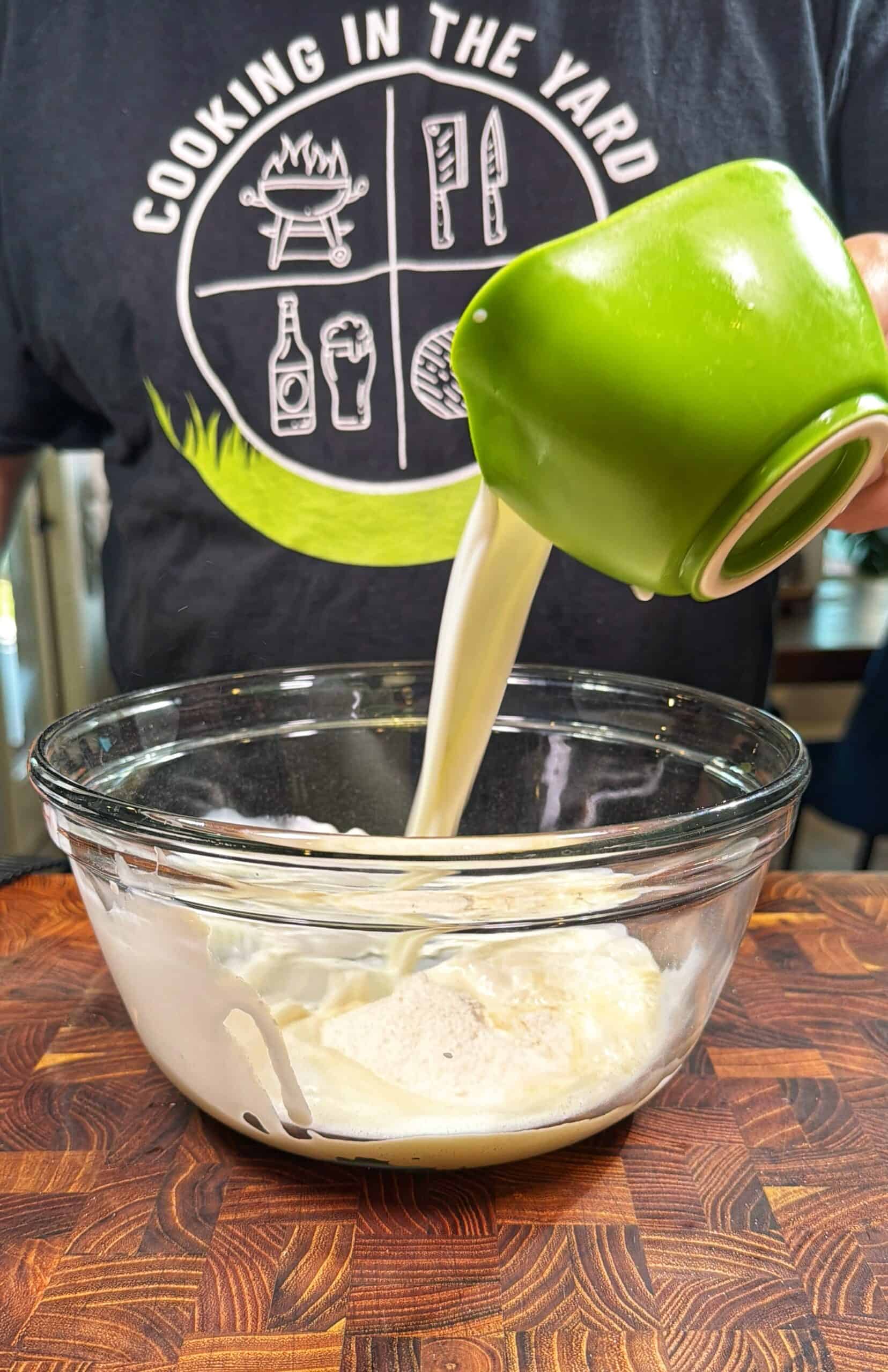 A person wearing a Cooking in the Yard shirt pours liquid from a green measuring cup into a glass bowl on a wooden surface. The bowl contains a creamy mixture.