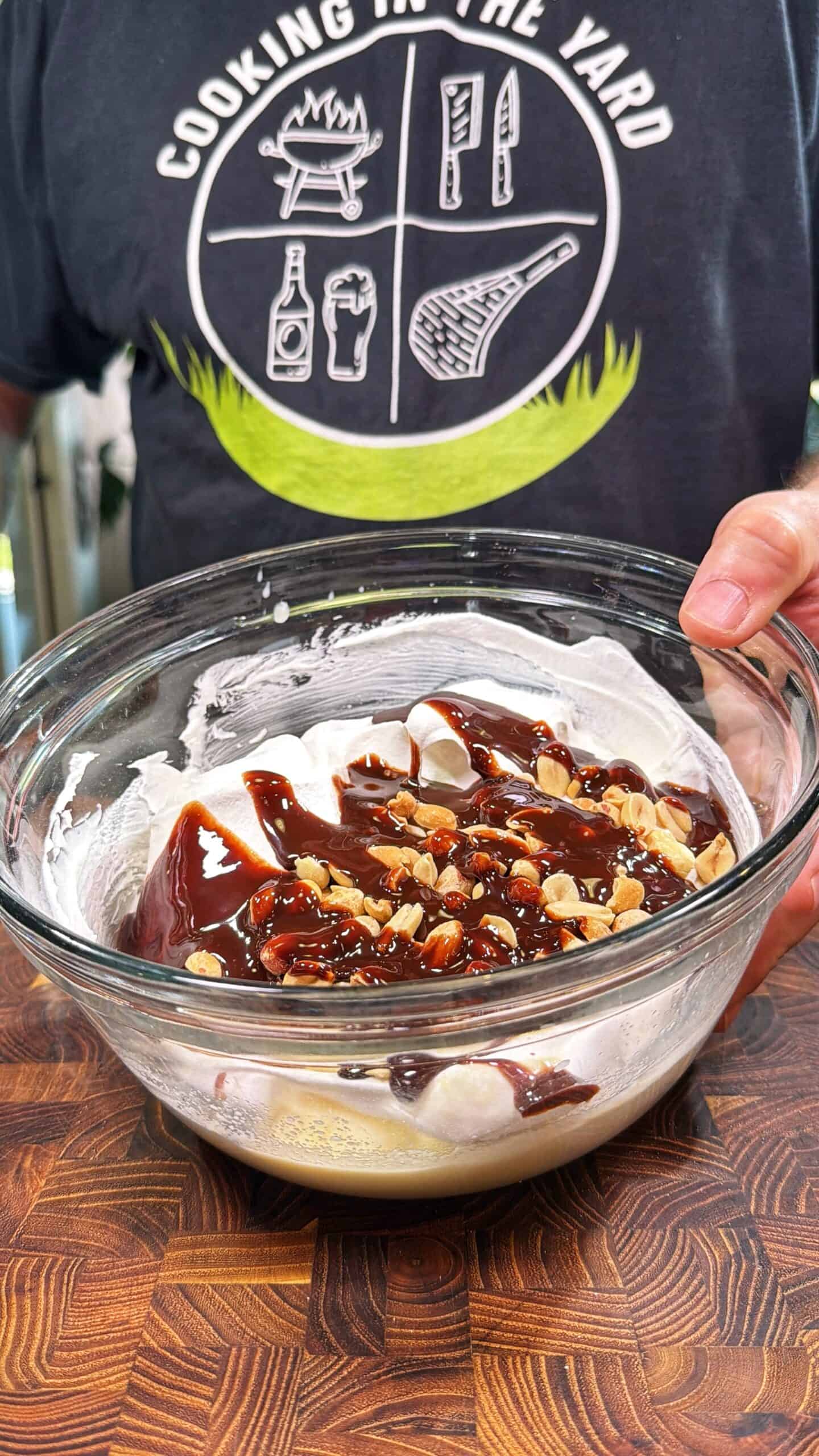 A person holding a glass bowl filled with marshmallows, chocolate sauce, and chopped nuts, standing over a wooden surface. The person is wearing a shirt that says Cooking in the Yard with barbecue-themed graphics.