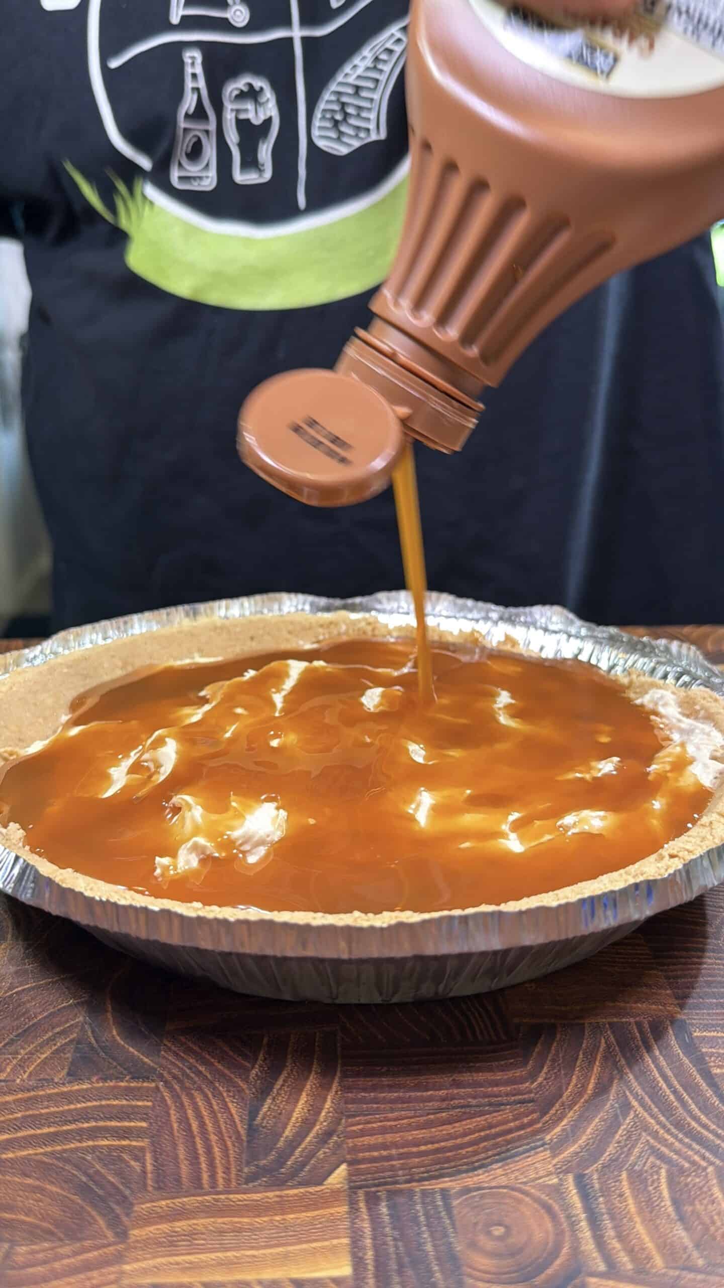 A person pours caramel sauce from a bottle onto a pie with a graham cracker crust. The pie sits on a wooden surface, and the person is wearing a black shirt with white graphics.