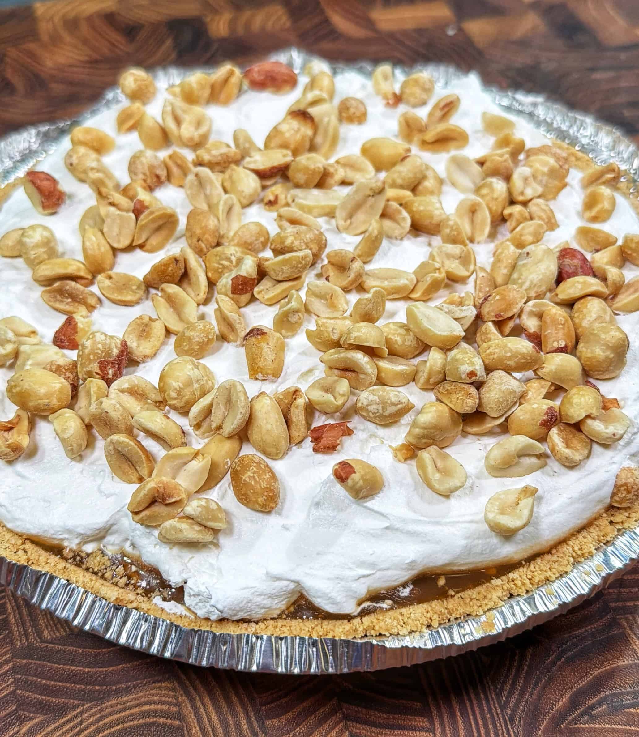 A pie in a foil tray topped with whipped cream and a generous layer of salted peanuts, set on a wooden surface.