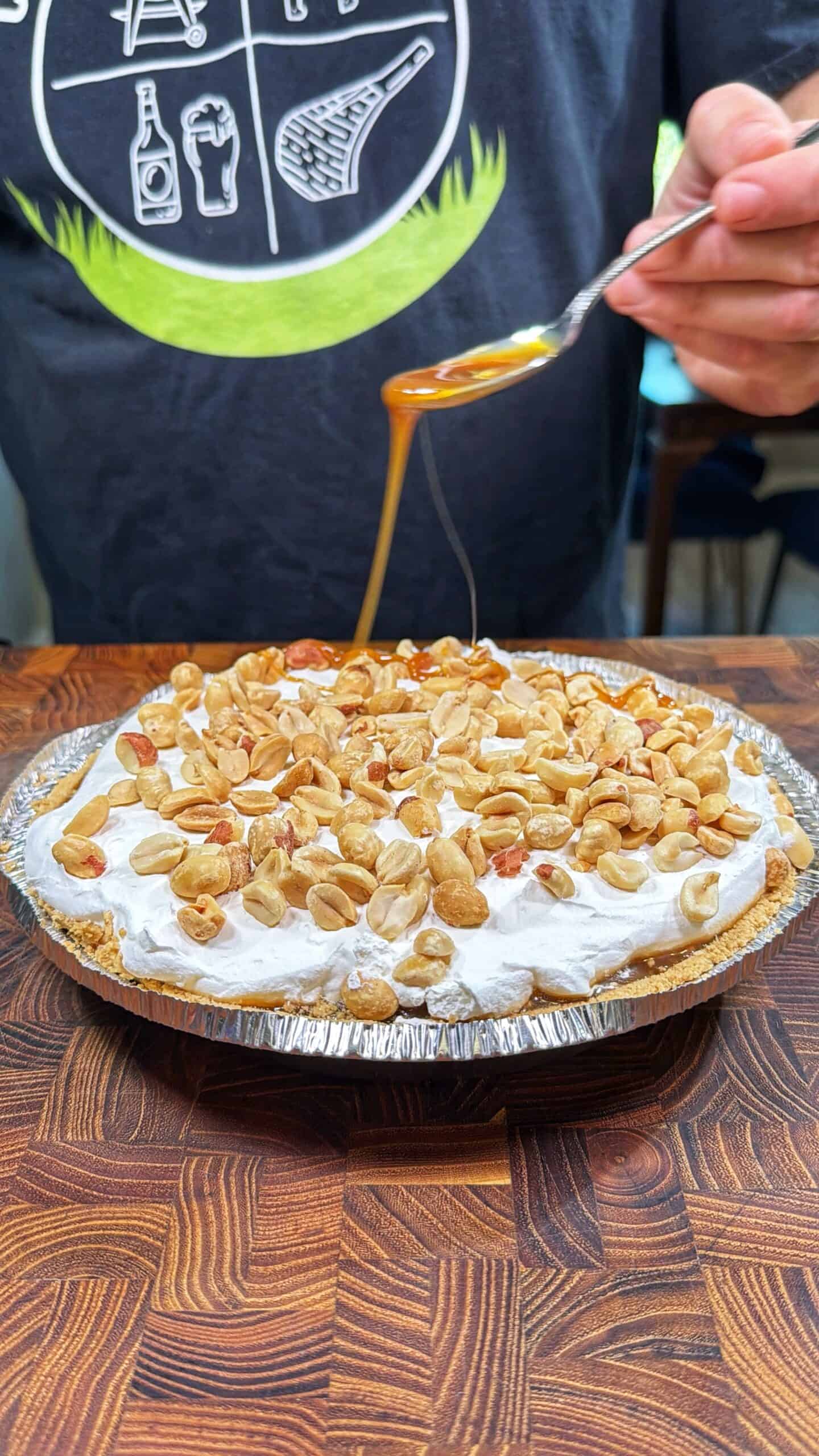 A person drizzles caramel sauce from a spoon onto a pie topped with whipped cream and peanuts, set on a wooden surface.