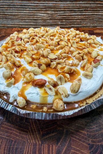 A pie topped with whipped cream, caramel sauce, and a generous layer of peanuts in a foil pie pan, set on a wooden surface with a rustic wood background.