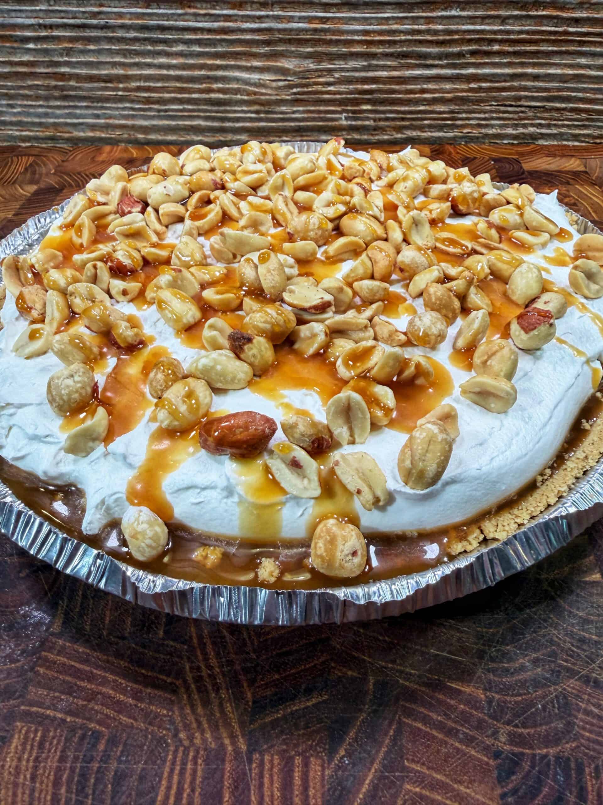 A pie topped with whipped cream, caramel sauce, and a generous layer of peanuts in a foil pie pan, set on a wooden surface with a rustic wood background.