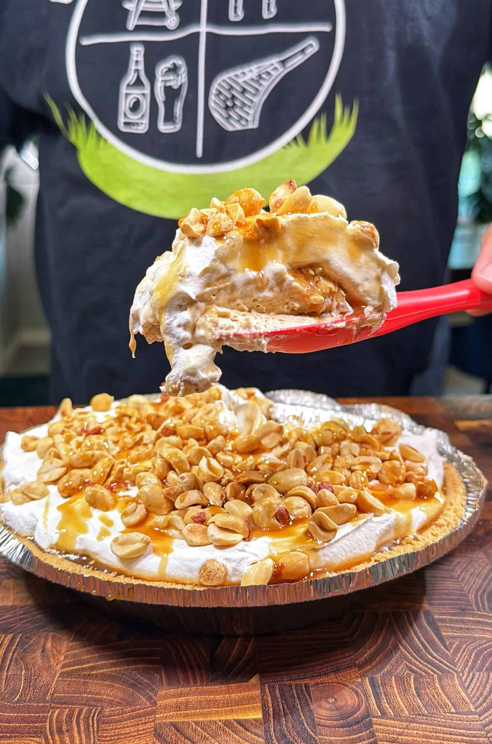 A close-up of a creamy pie topped with peanuts and caramel sauce, with a slice being lifted from the tin on a red spatula. A person in a dark shirt stands in the background.