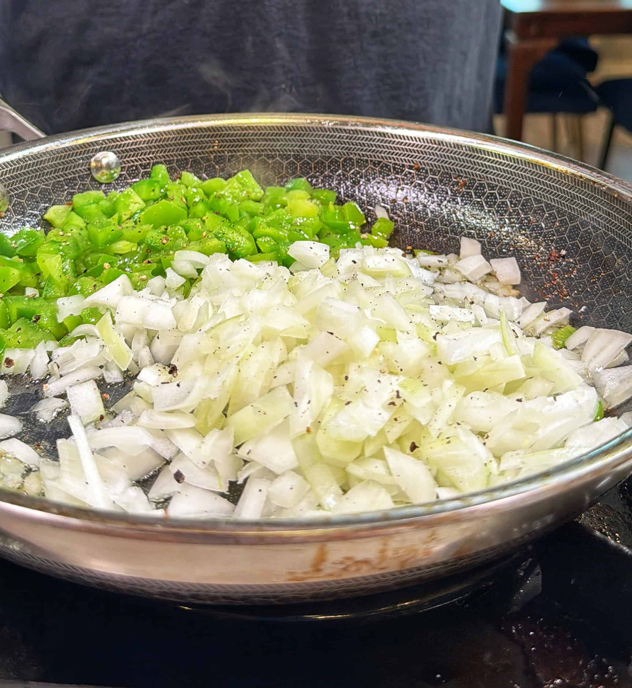 Chopped green bell peppers and onions are being sautéed in a stainless steel pan on a stovetop, with visible black pepper sprinkled on top.