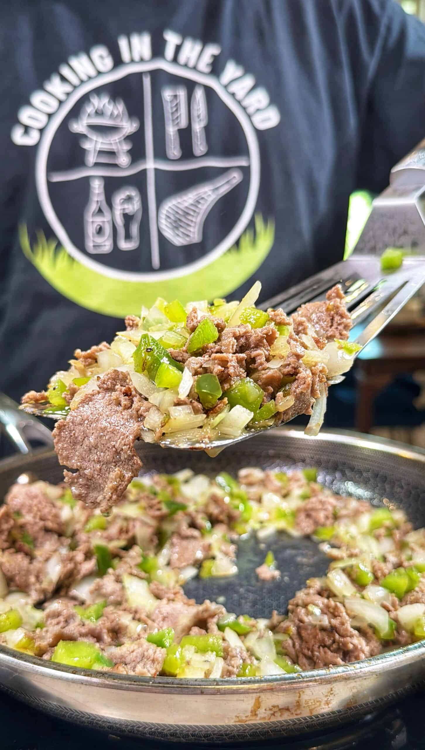 A close-up of cooked ground meat with diced green peppers and onions being scooped from a pan. In the background, a person wears a shirt that says COOKING IN THE YARD with food-themed graphics.