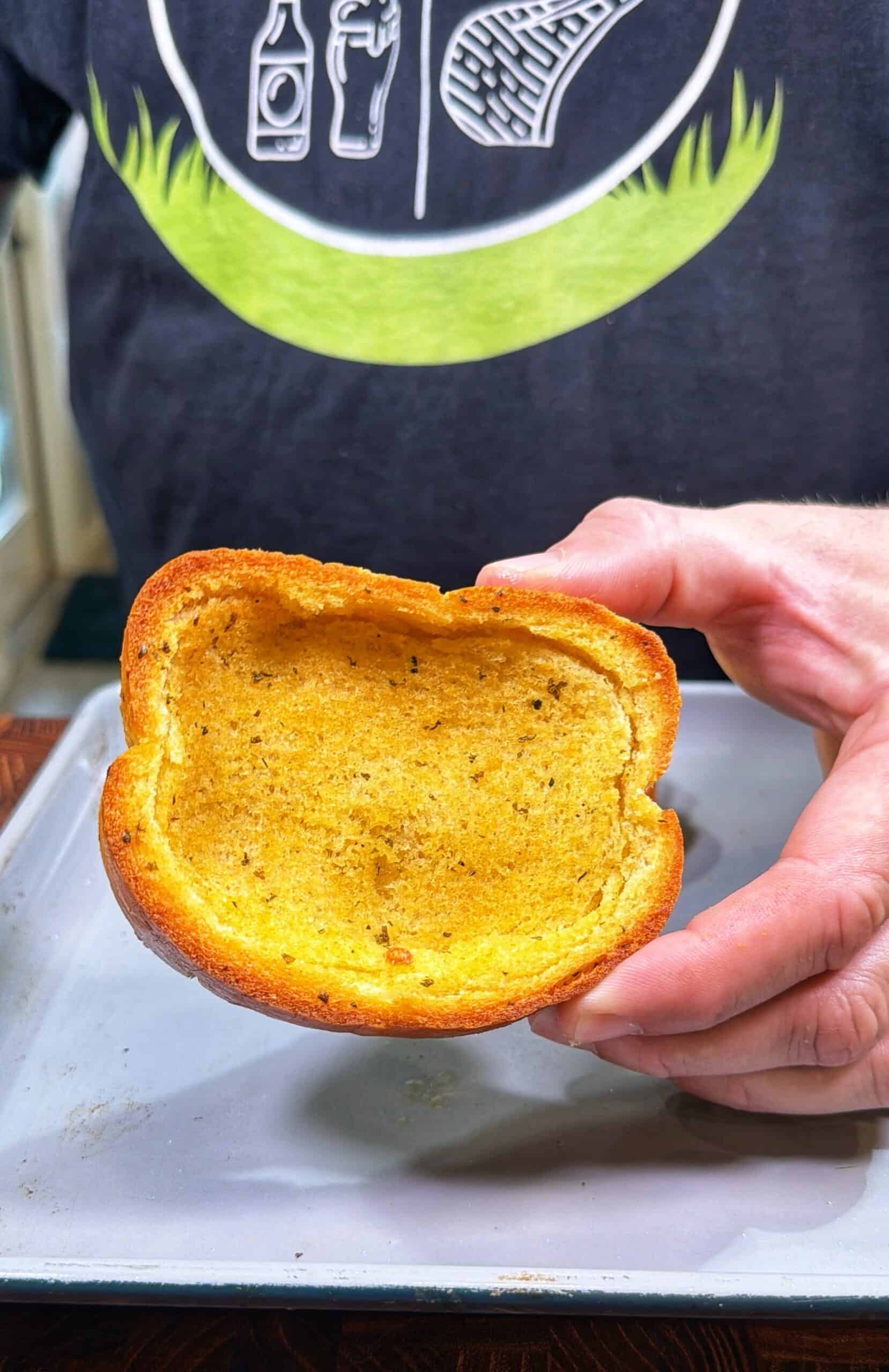 A person holds a large, toasted, hollowed-out piece of bread on a baking tray. The person is wearing a dark shirt with white and green graphics on it.
