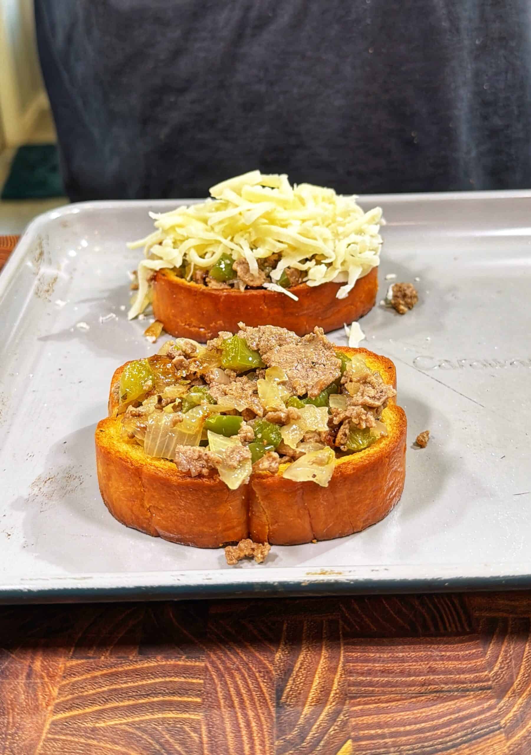 Two thick slices of bread on a baking sheet, each topped with cooked ground beef, onions, and green peppers; one slice also has a layer of shredded cheese on top.