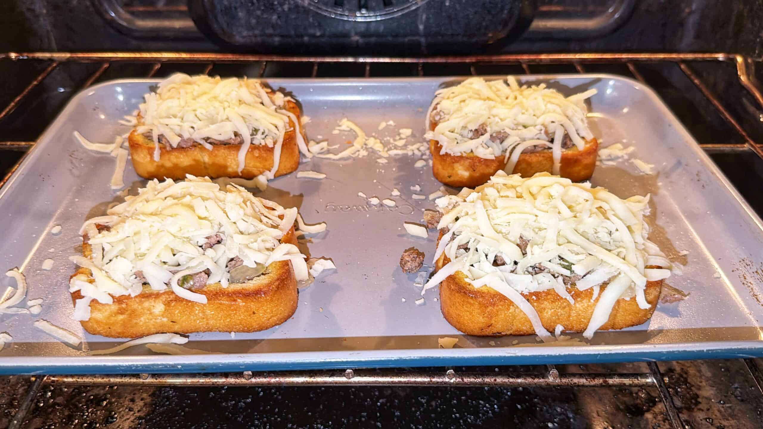 Four slices of bread topped with ground meat and shredded cheese are placed on a baking sheet inside an oven, ready to be baked.
