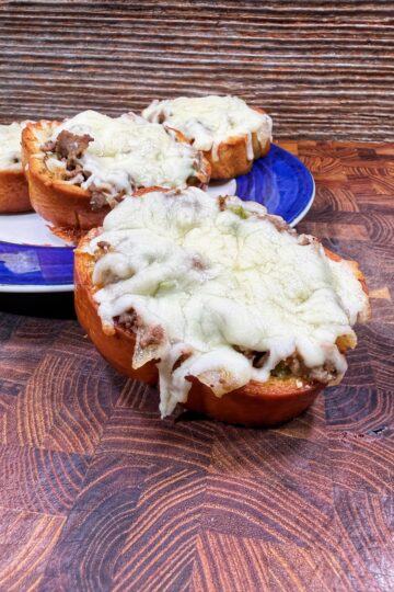 Three slices of bread topped with ground meat and melted cheese are arranged on a wooden surface, with two pieces on a blue-rimmed plate and one in front, all in front of a textured background.