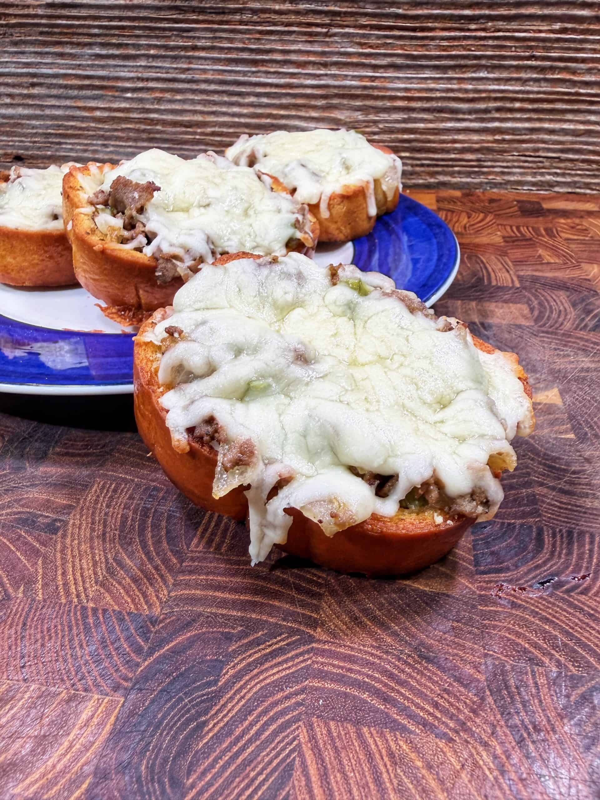 Three slices of bread topped with ground meat and melted cheese are arranged on a wooden surface, with two pieces on a blue-rimmed plate and one in front, all in front of a textured background.