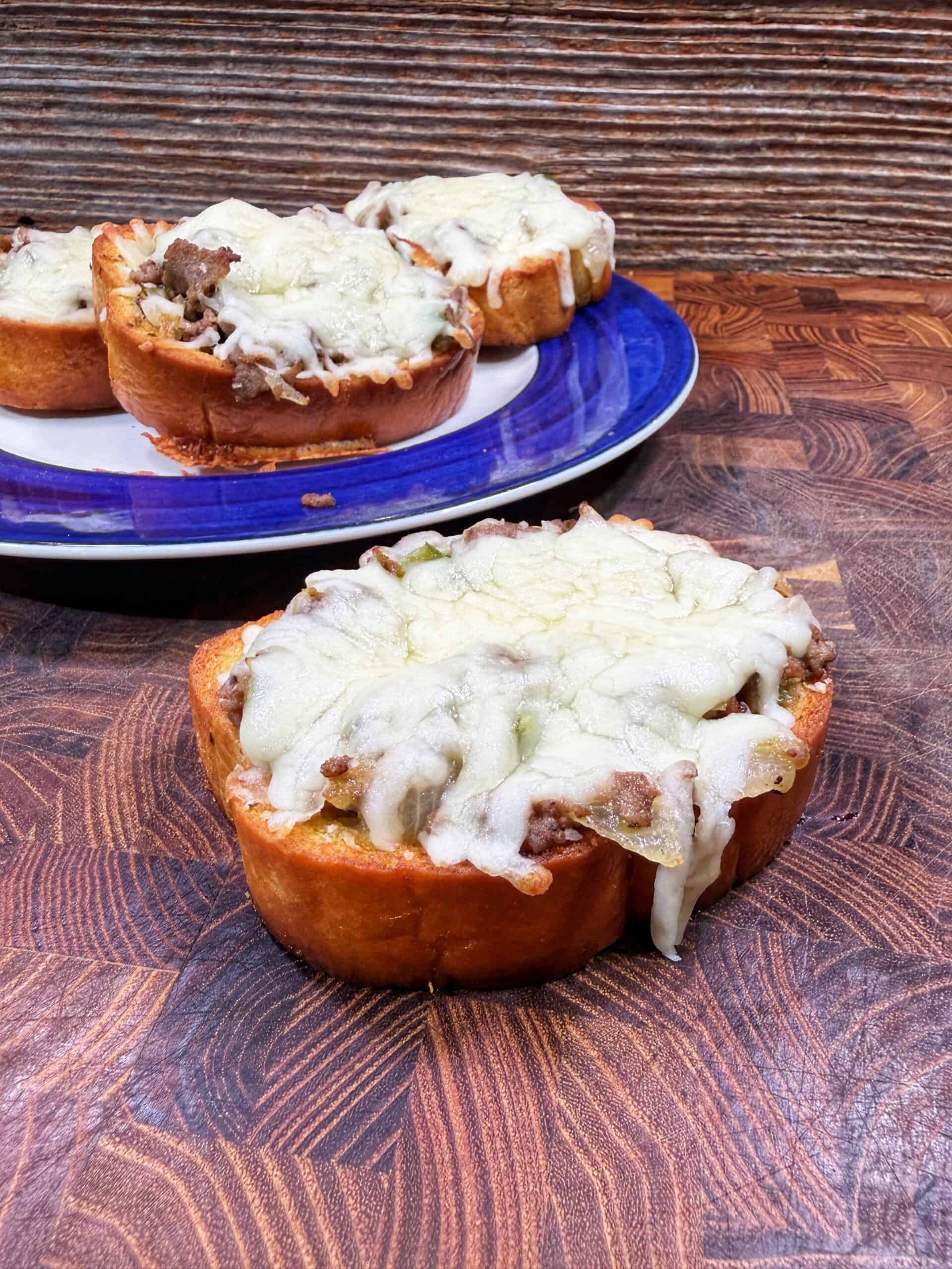 Thick slices of bread topped with ground meat and melted cheese are arranged on a wooden surface, with more of the same dish on a blue and white plate in the background.