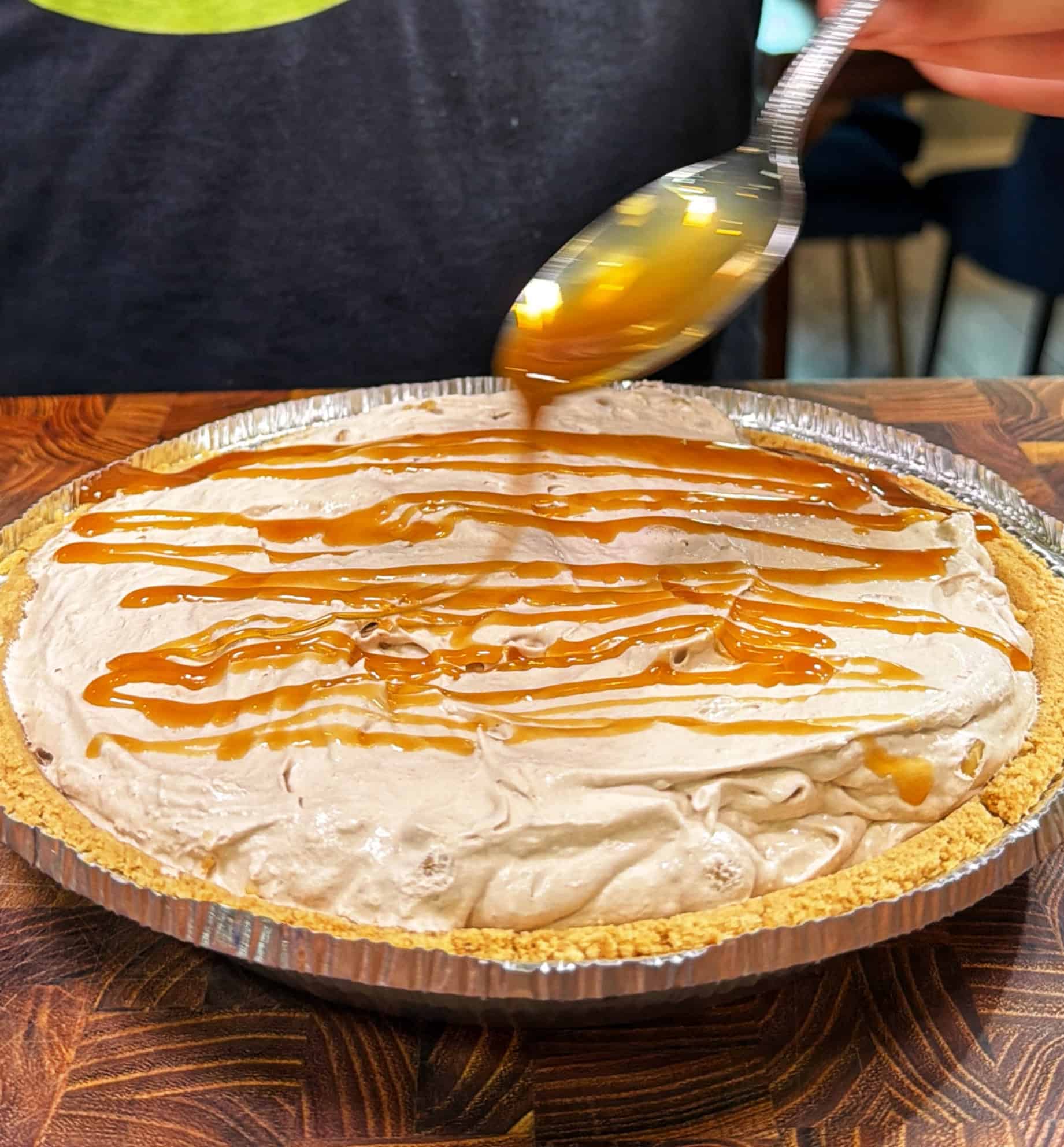 A person drizzles caramel sauce over a creamy pie with a graham cracker crust, using a spoon. The pie rests on a wooden surface.