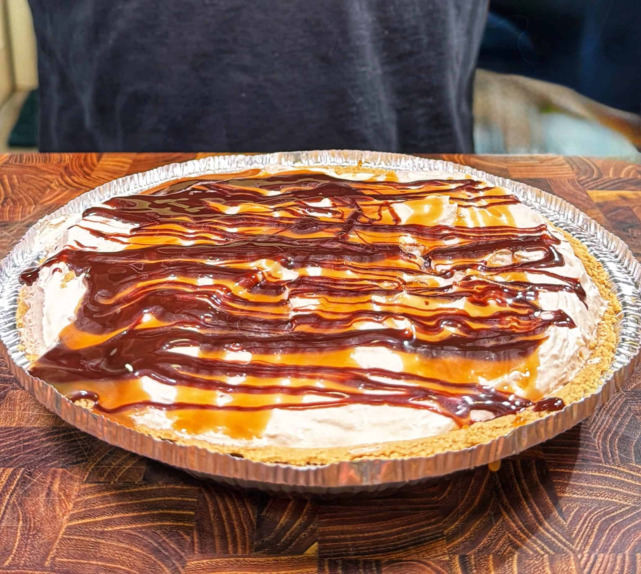 A pie in a foil pan sits on a wooden surface, topped with whipped cream and drizzled with caramel and chocolate sauces. A person in a dark shirt is partially visible behind the pie.