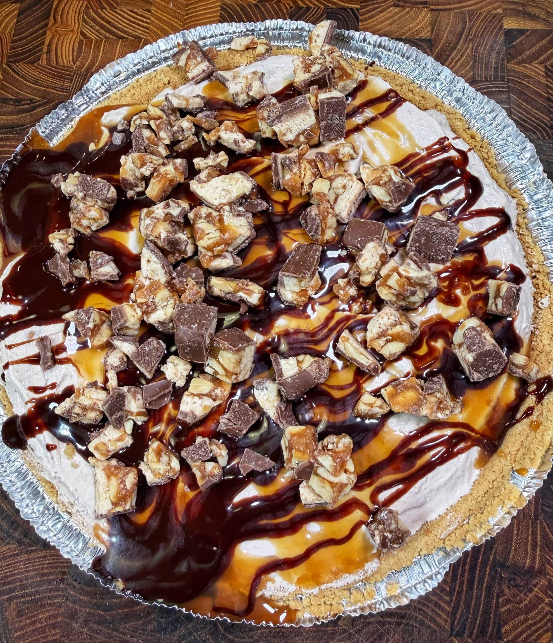 A pie with a graham cracker crust, topped with whipped filling, chopped chocolate candy bars, and drizzles of chocolate and caramel sauce.