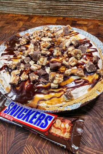 A pie with a crumb crust topped with chopped Snickers bars, chocolate, and caramel drizzle sits on a wooden surface next to an unopened Snickers candy bar.