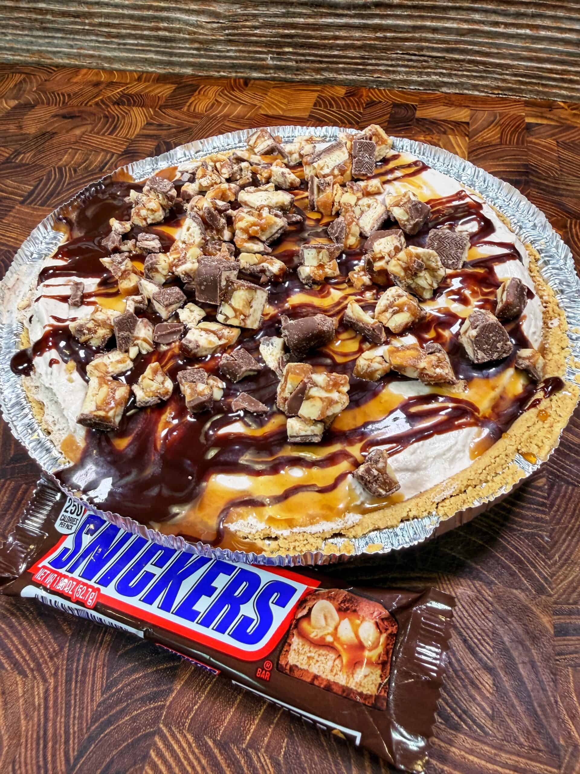 A pie with a crumb crust topped with chopped Snickers bars, chocolate, and caramel drizzle sits on a wooden surface next to an unopened Snickers candy bar.
