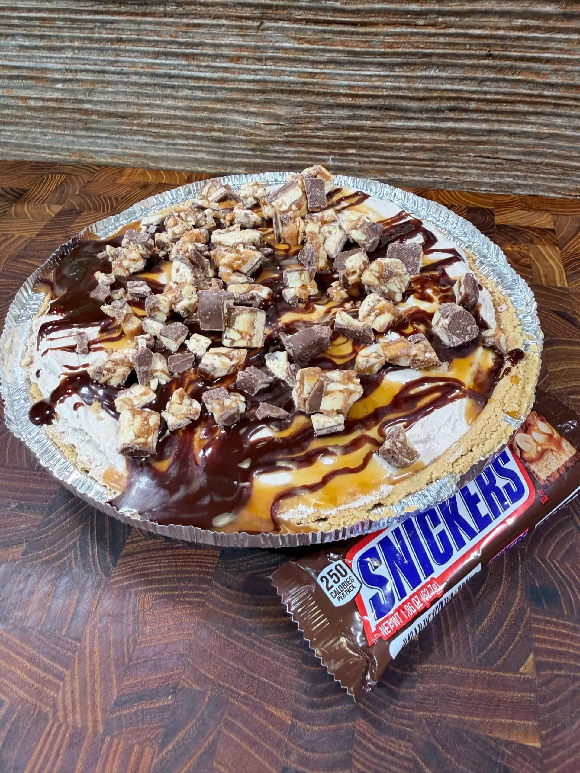 A pie topped with chopped Snickers bars, drizzled with chocolate and caramel sauce, sits on a wooden surface next to an unopened Snickers candy bar.