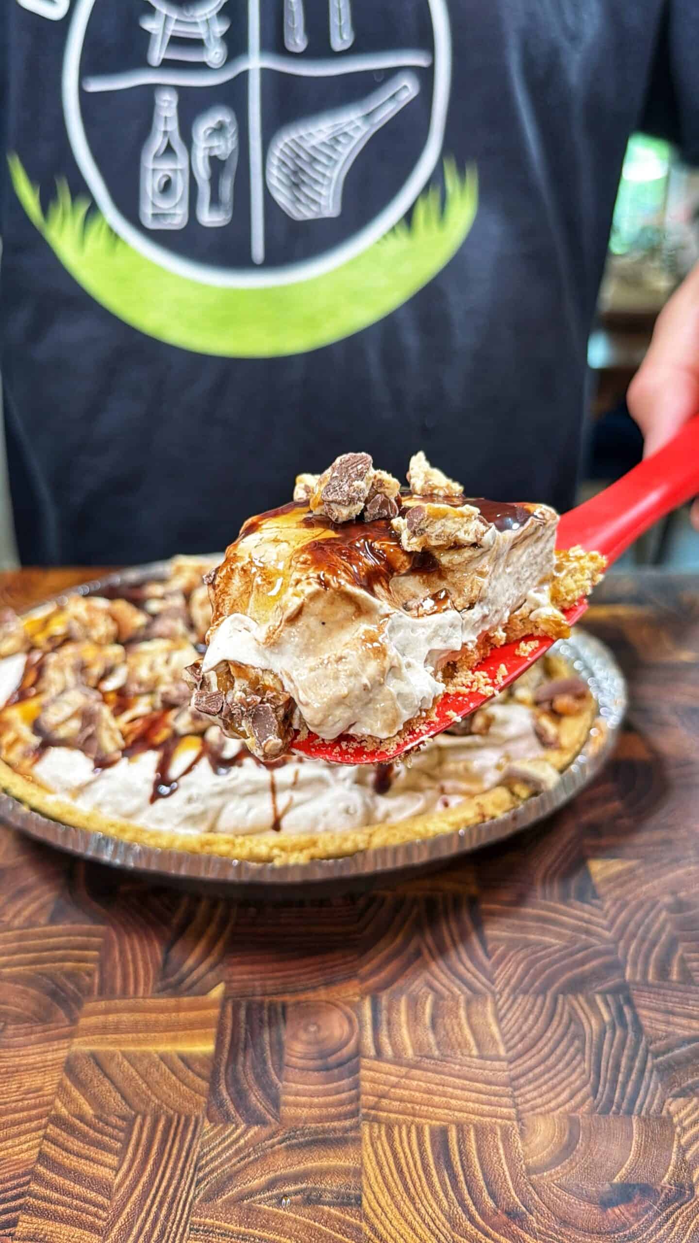 A close-up of a pie slice on a red spatula, topped with chocolate syrup and chopped candy, held above the rest of the pie on a wooden surface. A person in a dark shirt with white graphics is in the background.