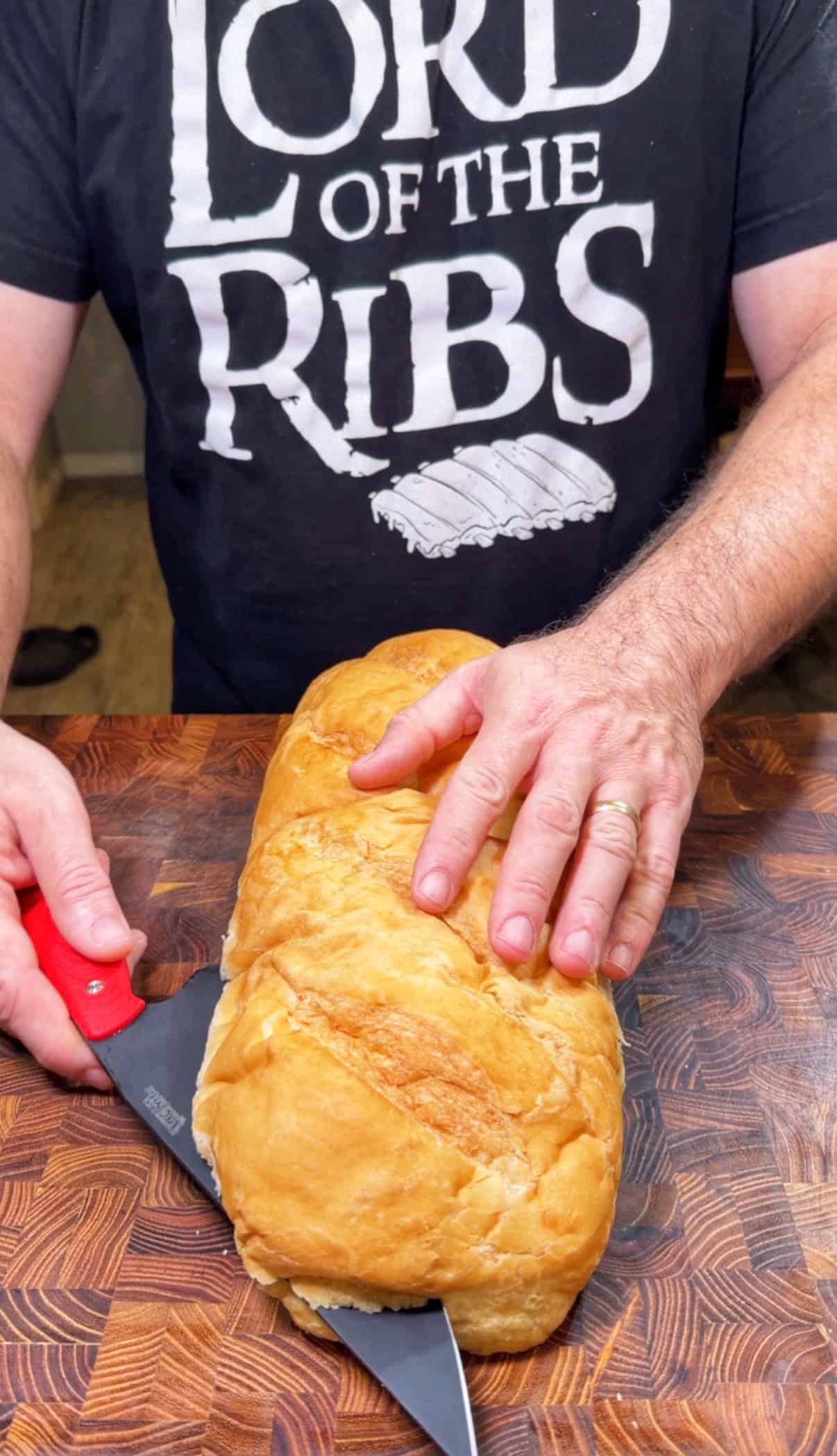 A person wearing a Lord of the Ribs shirt is slicing a loaf of bread on a wooden cutting board with a red-handled knife.
