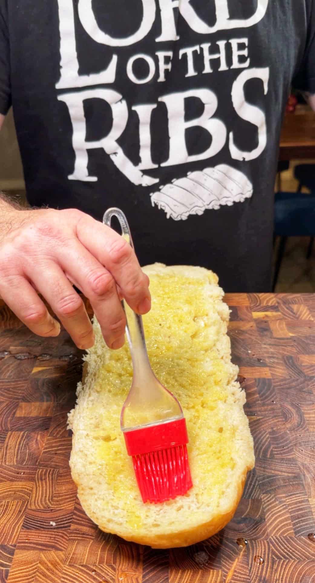 A person wearing a Lord of the Ribs shirt brushes melted butter onto a sliced loaf of bread on a wooden cutting board.